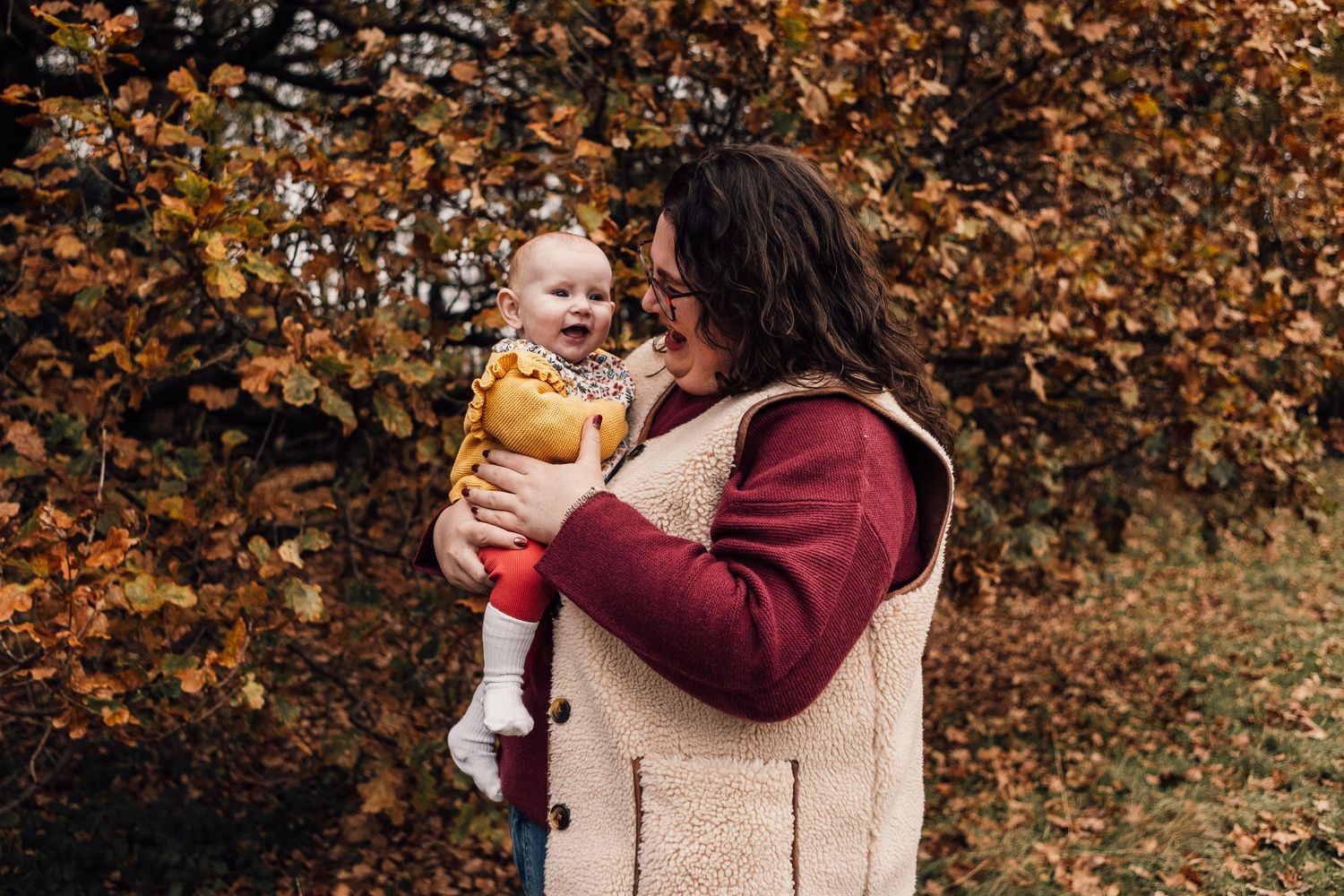 A tender autumn moment captured outdoors with fall foliage in the background, featuring warm burgundy and mustard colors.