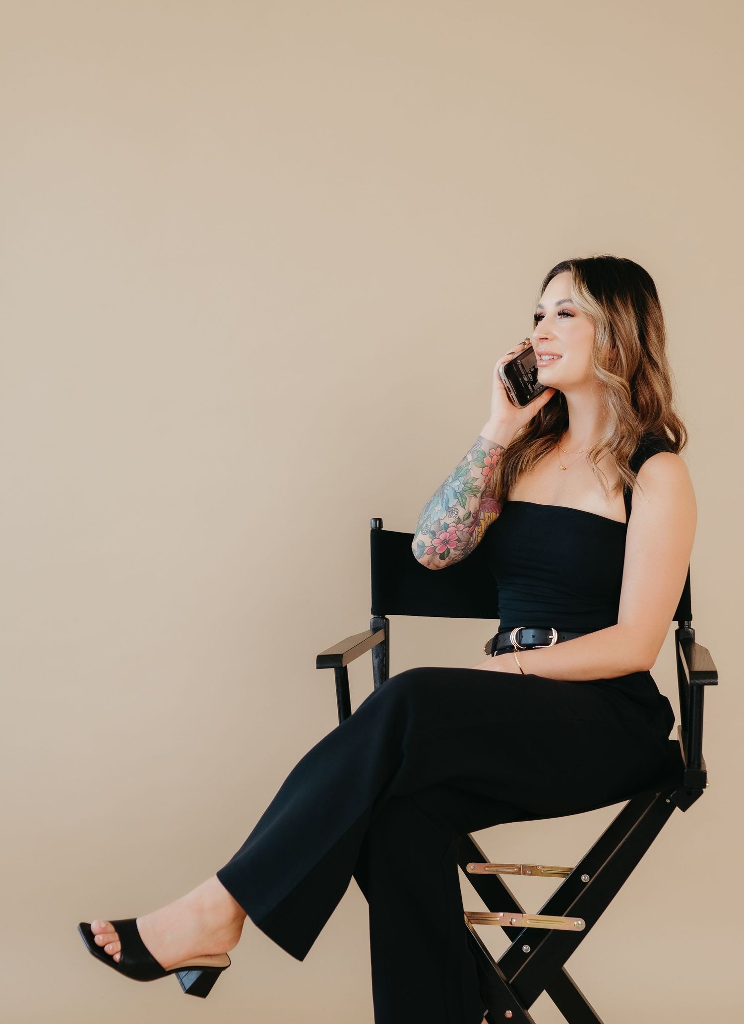 Woman in black outfit sitting in director's chair while talking on phone.