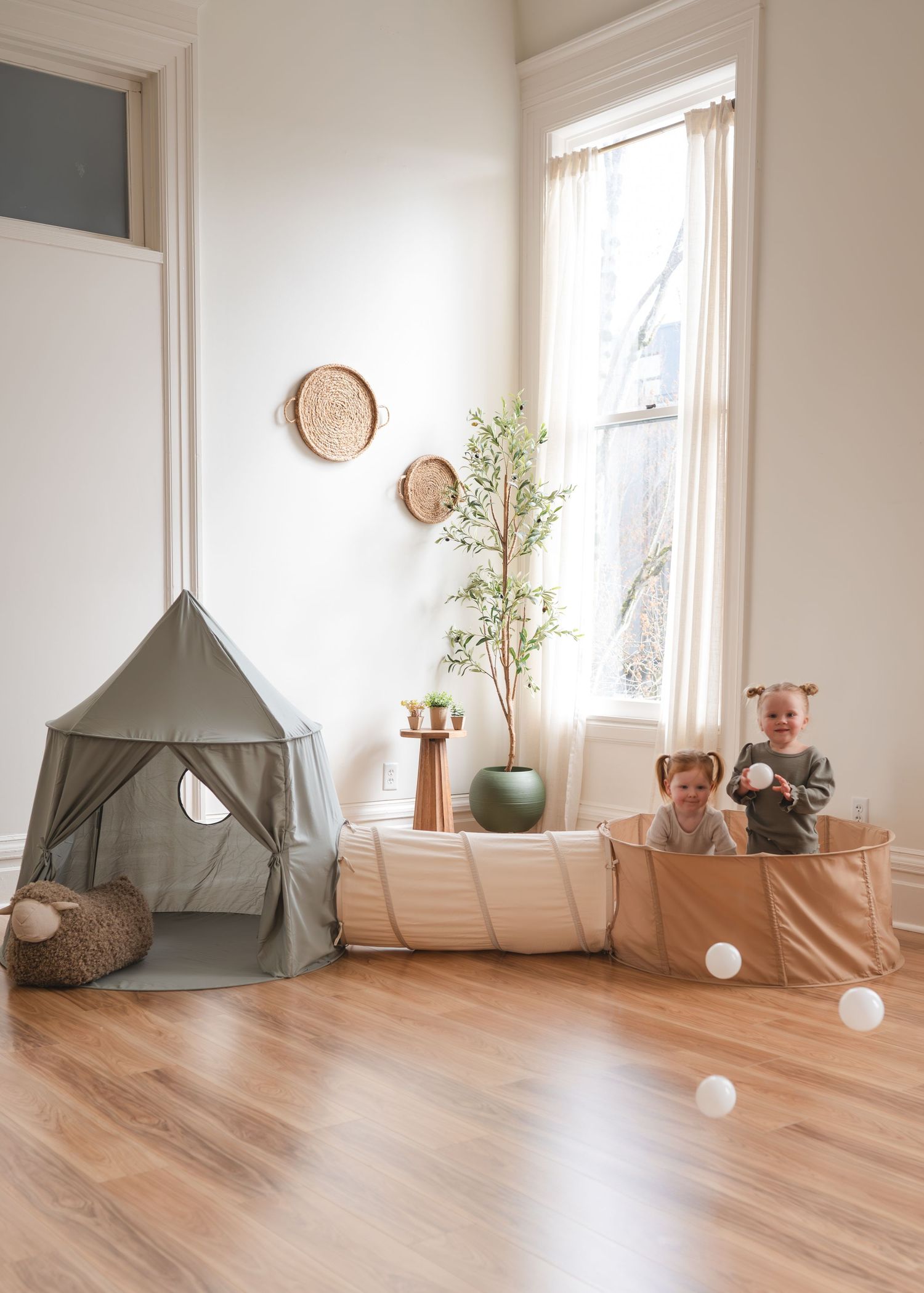 Simple children's playroom features a gray teepee tent, houseplants, and white walls with hardwood flooring.