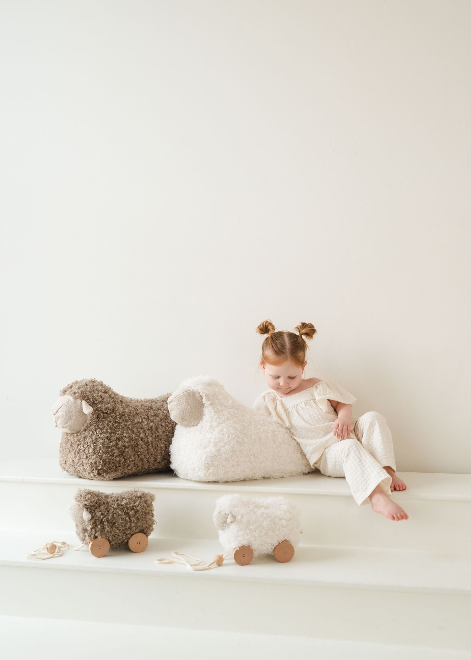 Small child in white outfit sits on bench with plush lamb toys in minimalist neutral room setting.