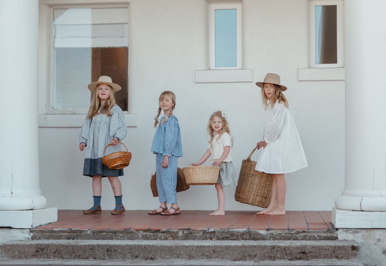 Four people in vintage-style clothing stand on a white porch with pillars while holding woven baskets.