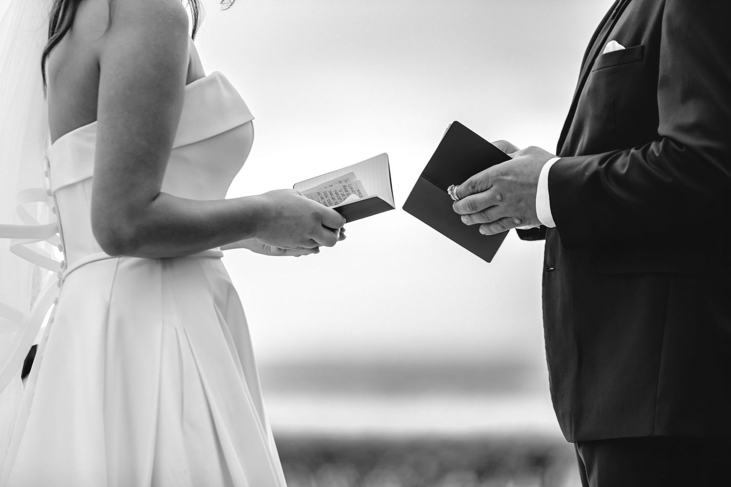 A bride and groom exchange vows while holding papers during their black and white outdoor wedding ceremony.