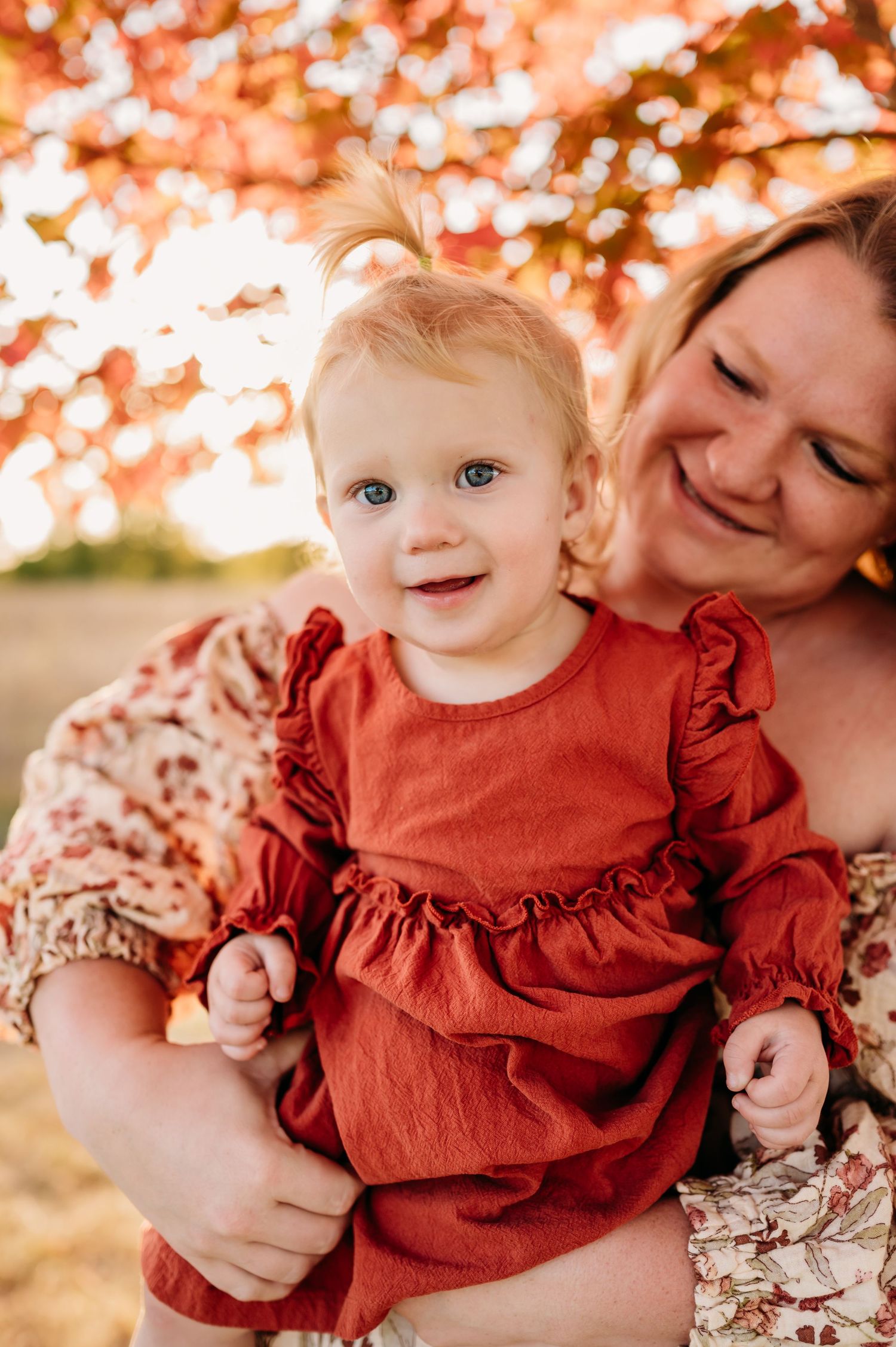 A toddler in a red ruffled dress smiles while being held outdoors during autumn with warm bokeh lighting in background.