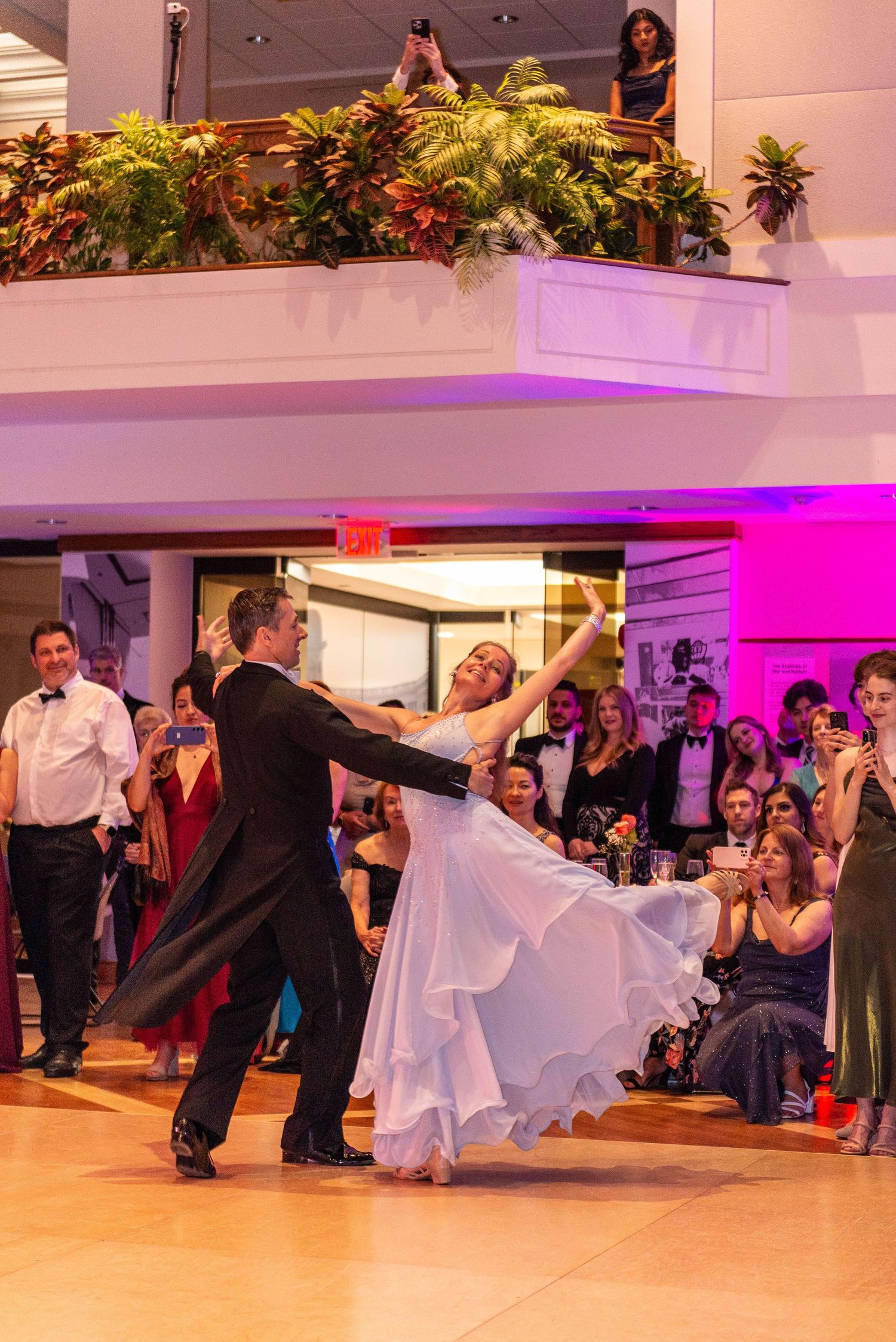 Elegant couple performing romantic ballroom dance at wedding reception with pink and purple lighting.