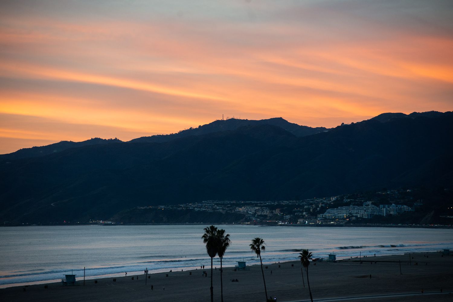 Coastal mountain silhouette against vibrant orange sunset sky with palm trees in foreground.