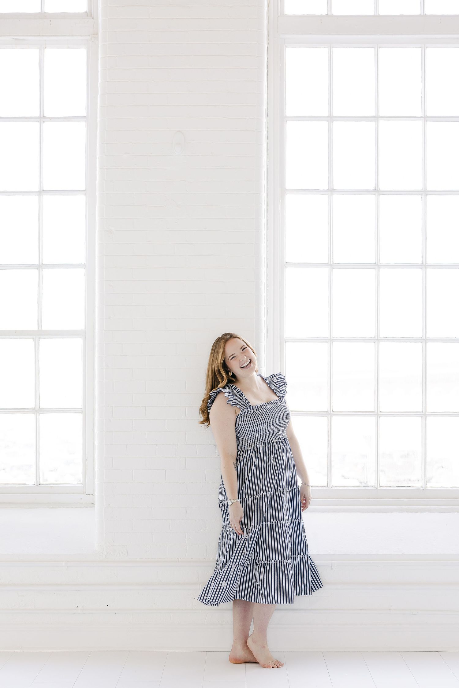 A joyful figure in a navy striped ruffle dress poses gracefully against white walls and windows in a bright minimal space.