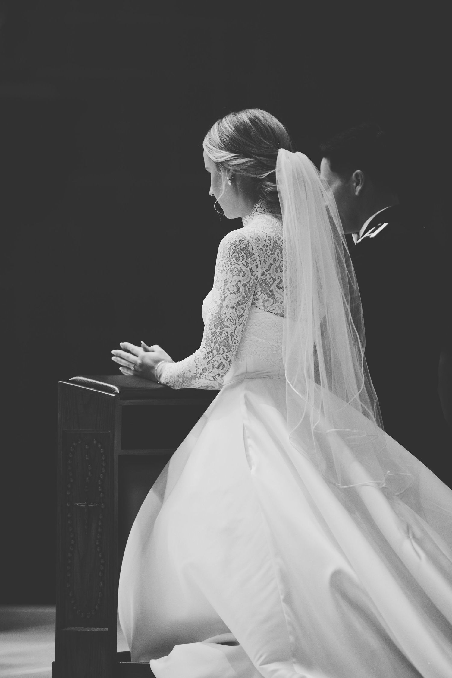 Bride in elegant lace gown and flowing veil kneels in prayer during wedding ceremony.