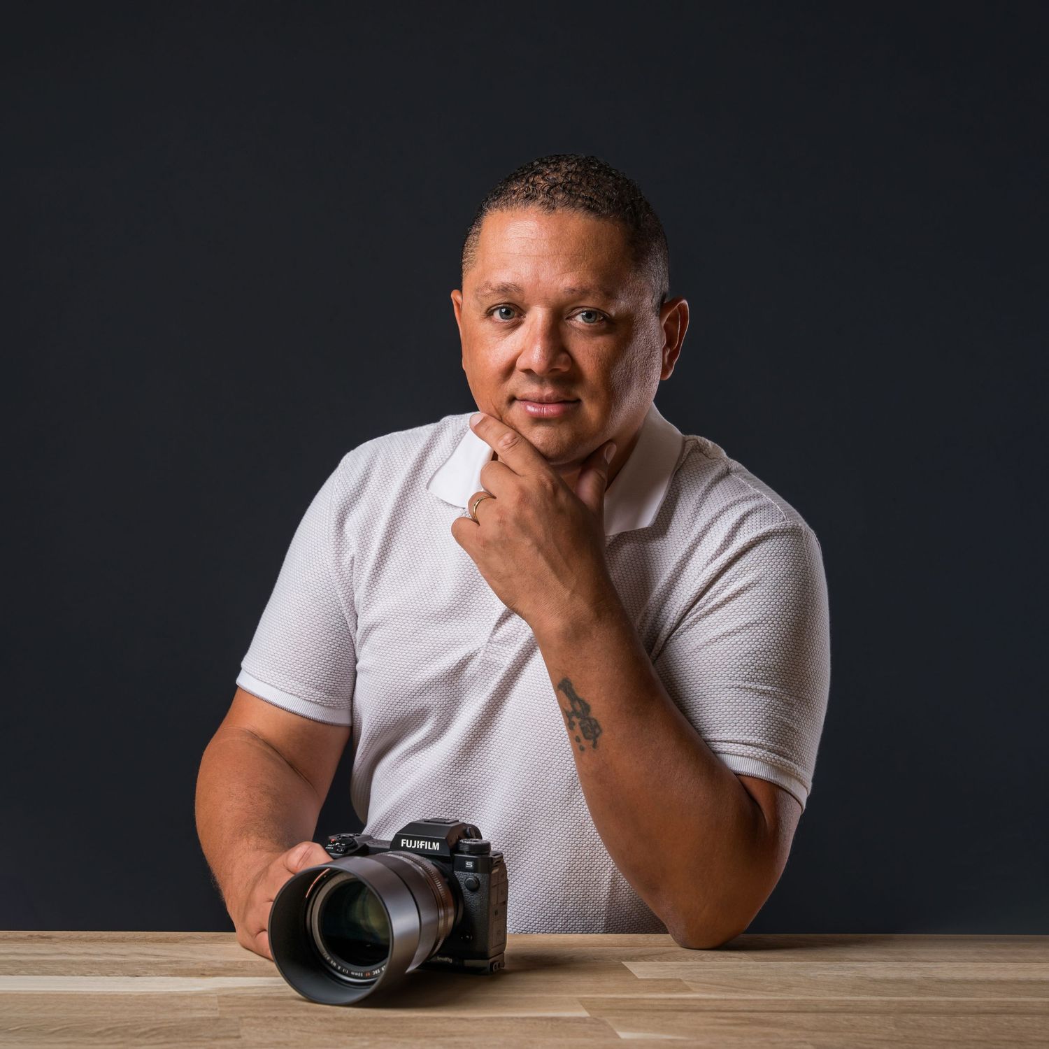 A photographer with a professional camera sits thoughtfully at a wooden table against a dark background.