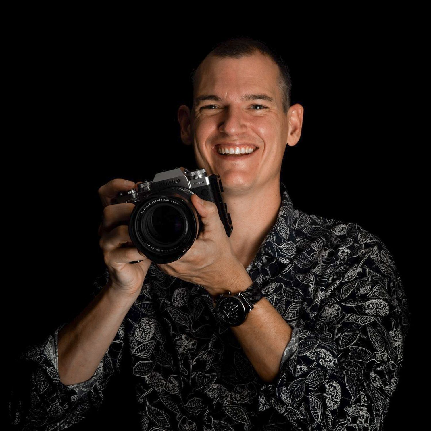 Photographer in dark floral shirt holding professional camera against black background.