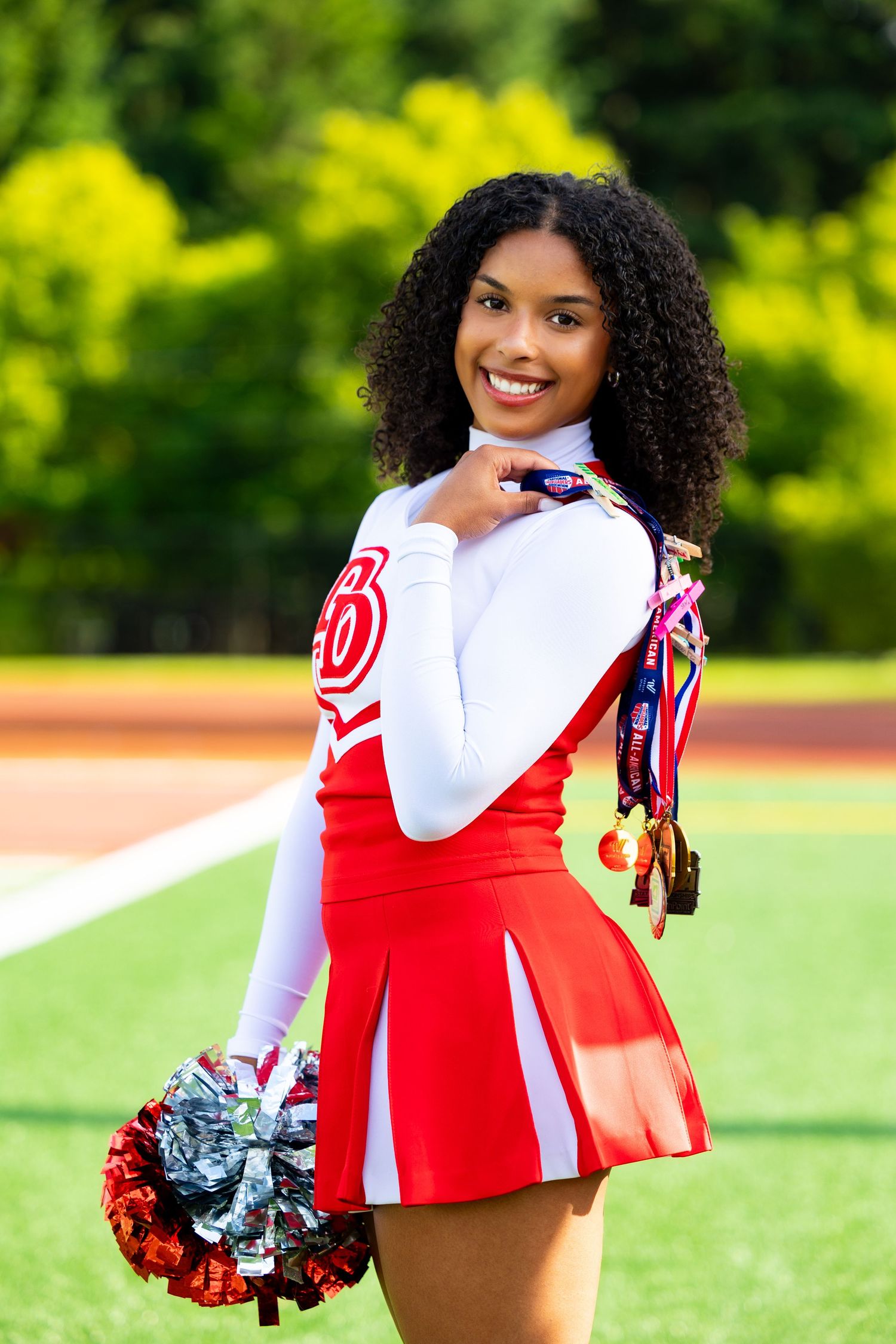 A cheerleader in a red and white uniform holds pom-poms while posing on an athletic field with yellow flowers in background.
