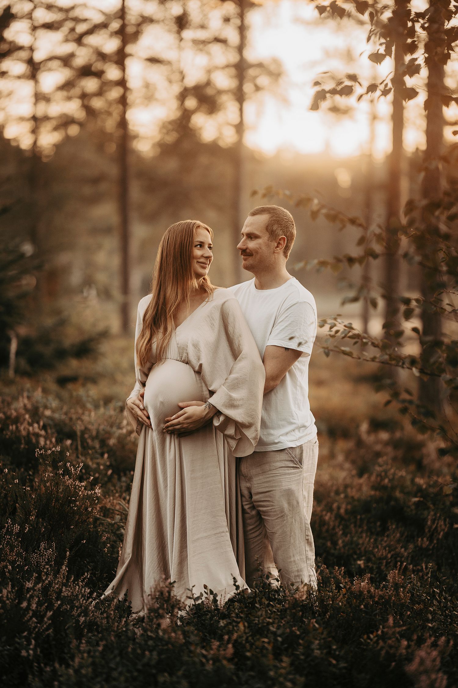 Expecting couple shares an intimate moment in a sunlit forest during maternity photoshoot.