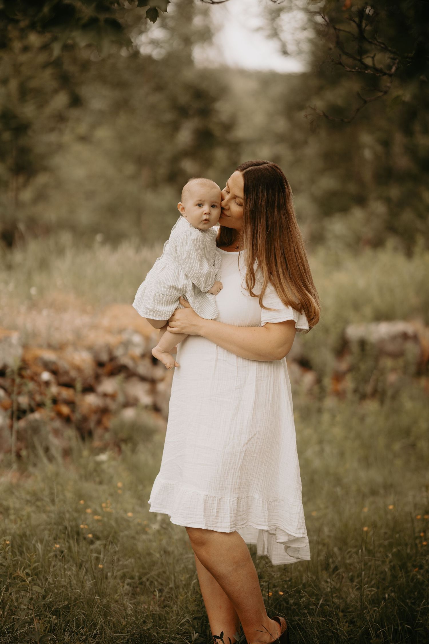 Mother and baby in matching white dresses stand together in a dreamy outdoor setting with bokeh background.