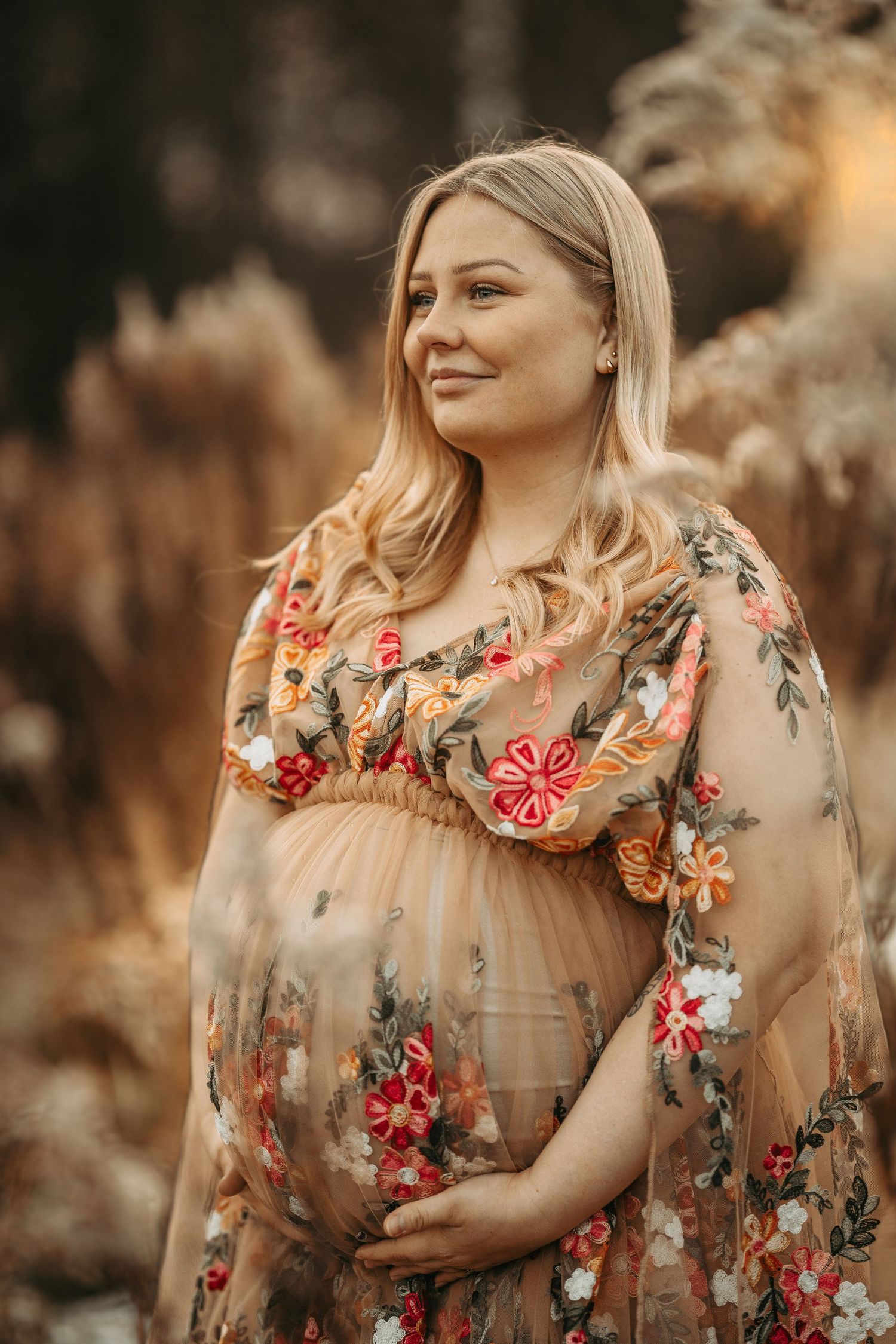 Expecting mother wearing floral dress poses outdoors during golden hour in autumn field.
