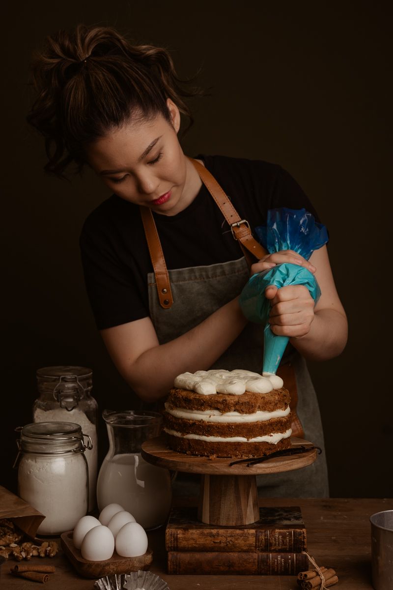 Baker using blue pastry bag to pipe frosting onto layered chocolate cake with baking ingredients on rustic wooden table.
