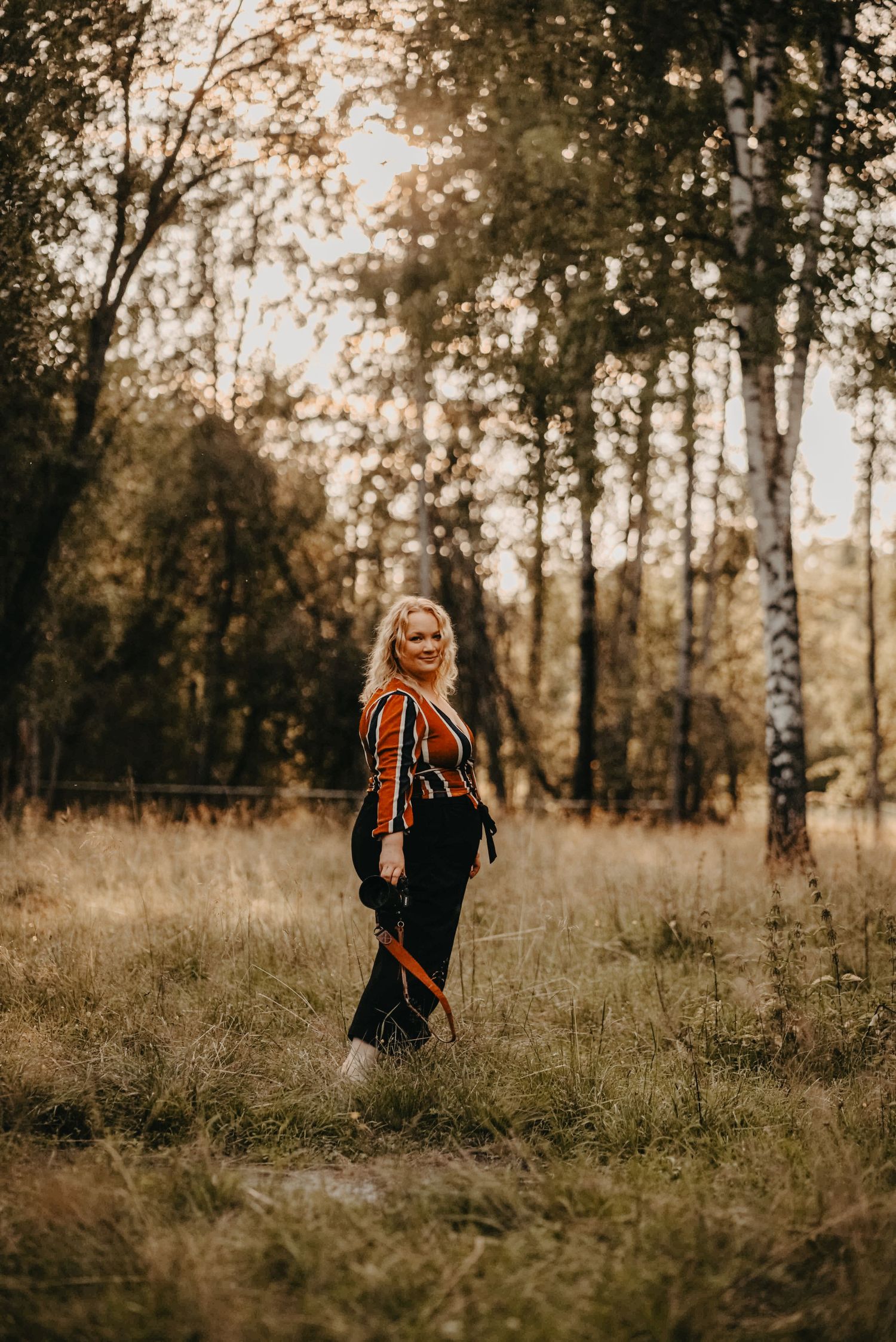 A person in a striped top and dark skirt stands in a sunlit autumn forest with golden light filtering through trees.