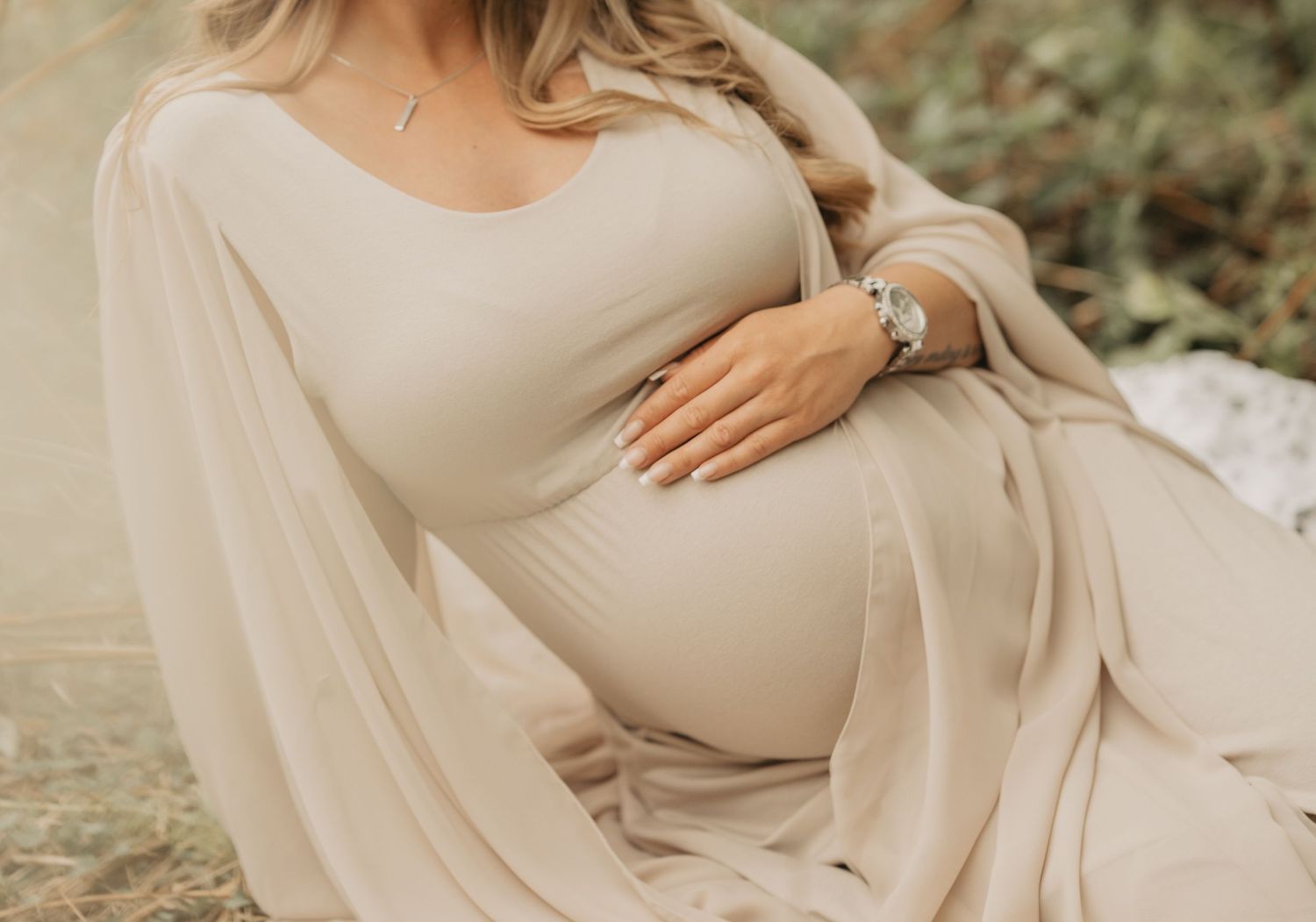 Pregnant woman in beige maternity dress and flowy shawl caresses her baby bump while sitting outdoors.