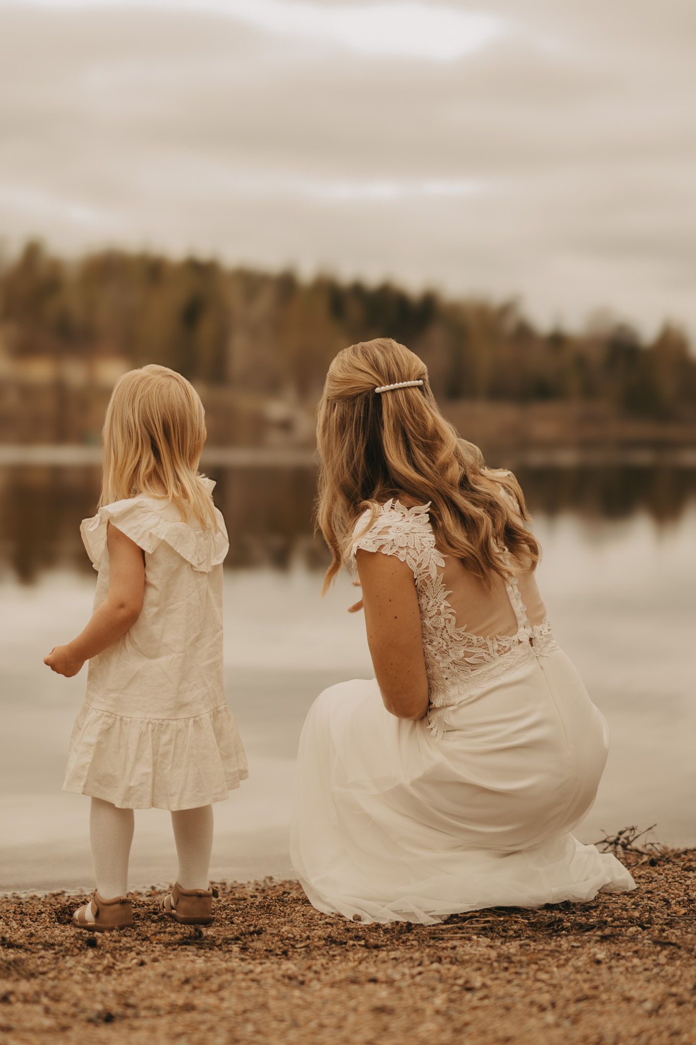 Woman and child in white dresses sit by tranquil lake at dusk with autumn trees in background.
