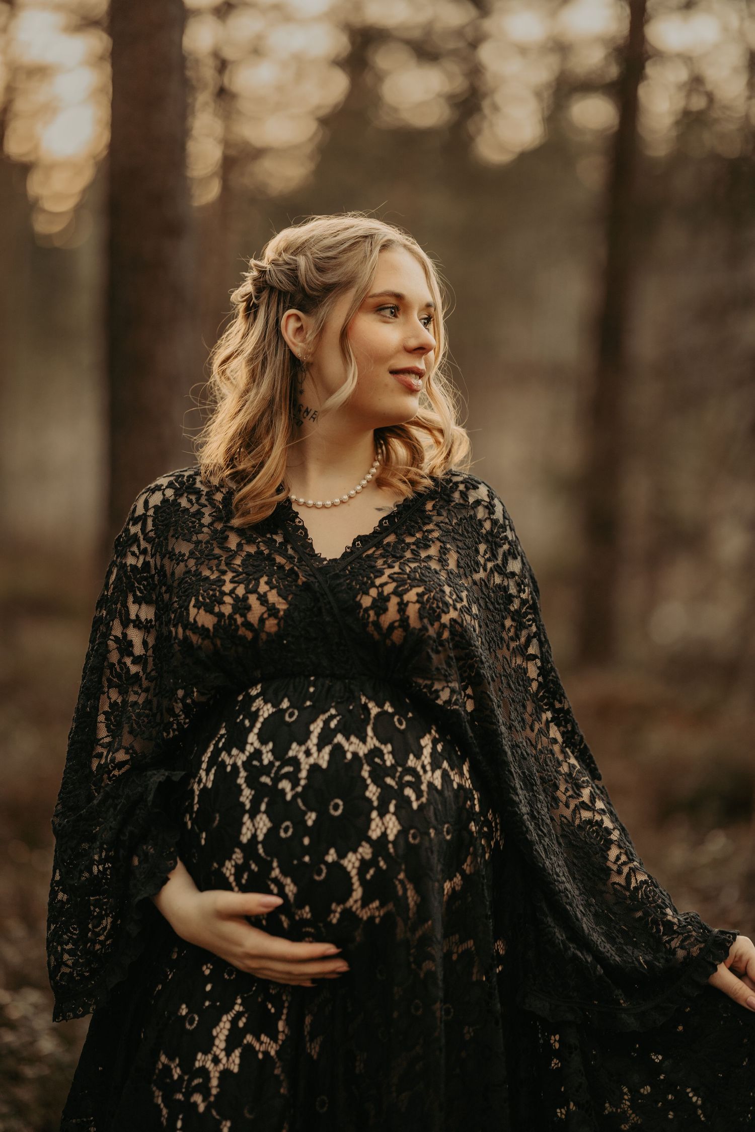 Ethereal maternity portrait in black lace dress against golden bokeh forest background at sunset.