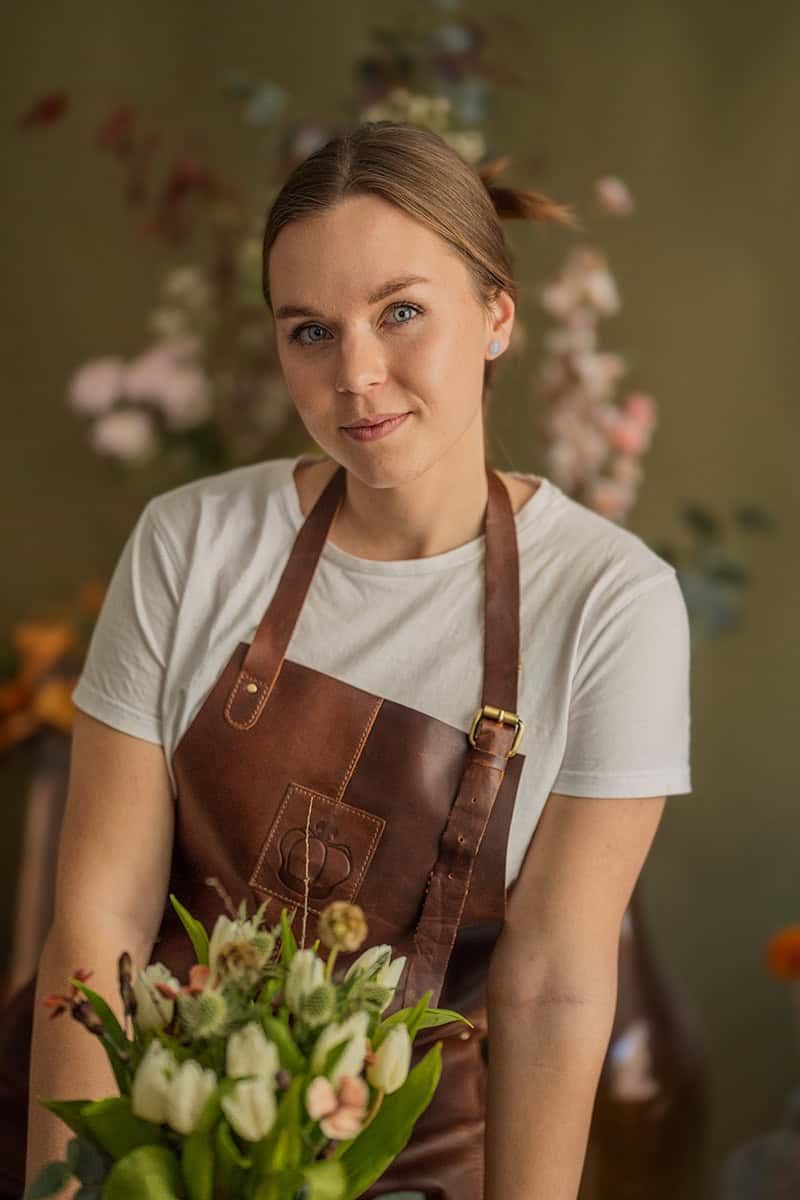 A florist wearing a brown leather apron holds fresh tulips in a warm-lit studio.