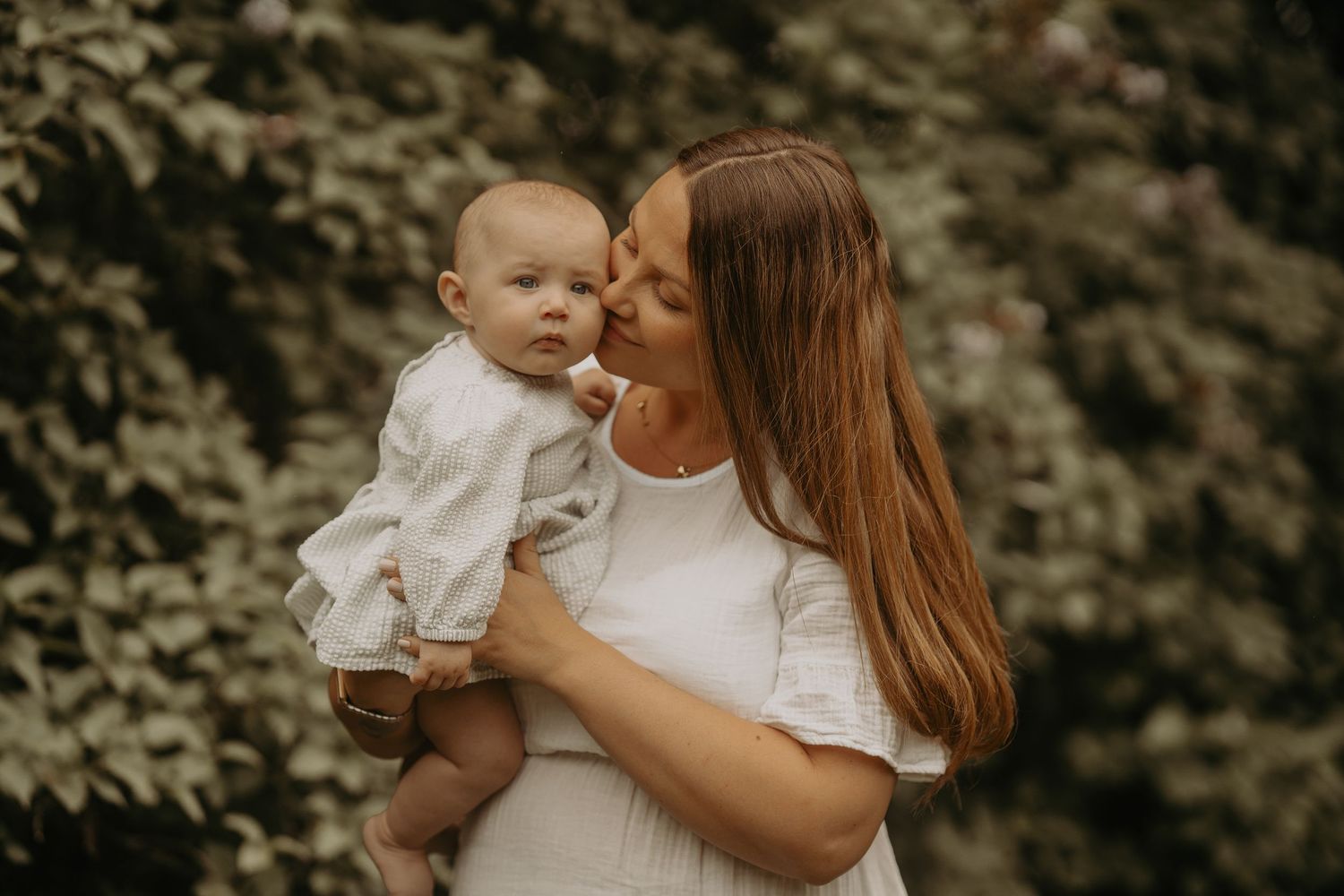 Woman in white dress holds and kisses child outdoors in front of lush green foliage backdrop.