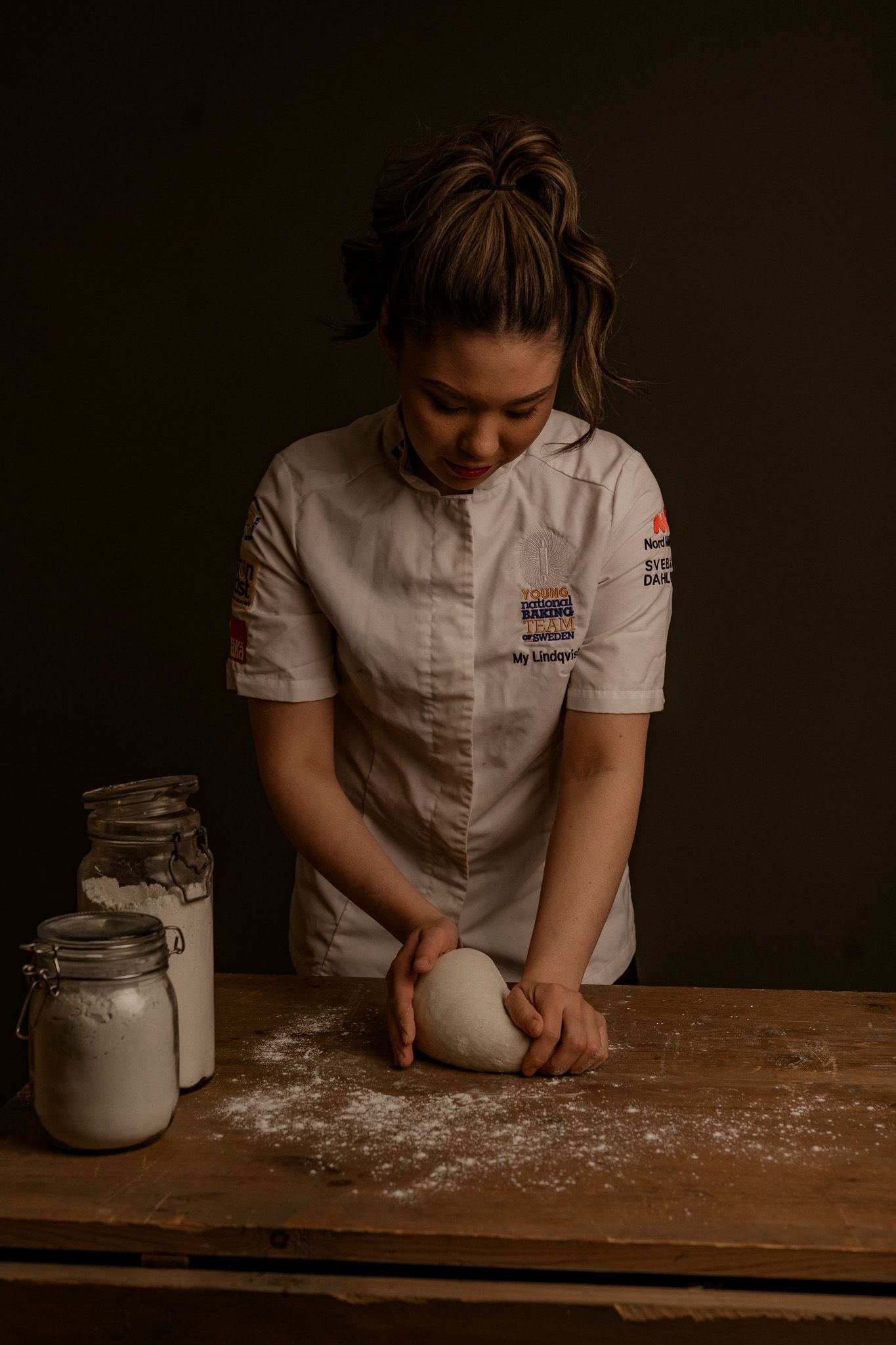 Chef in white uniform kneads dough on wooden table with flour and glass jars in moody lighting.