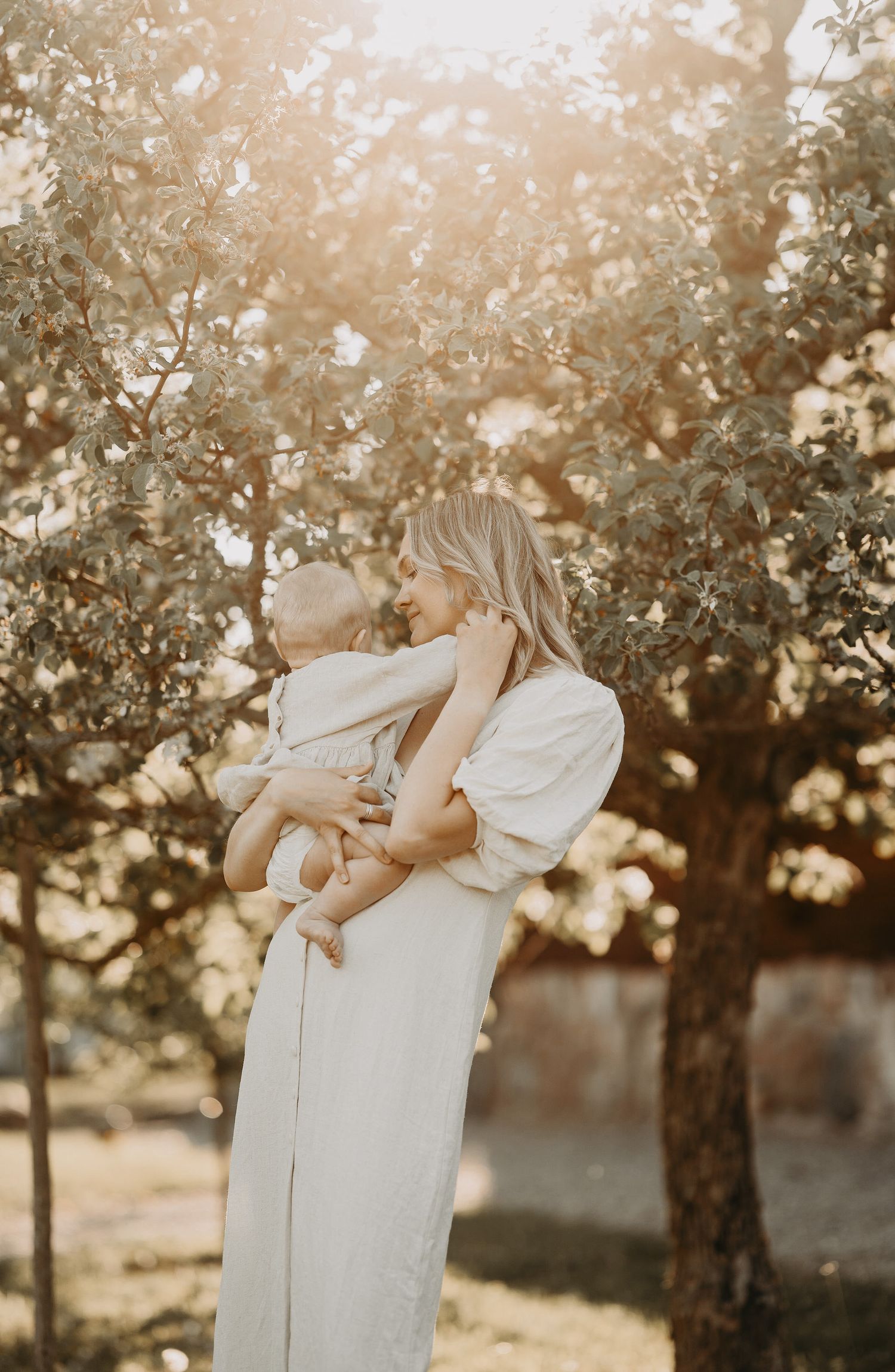 A tender outdoor moment captured in warm sunlight with trees and dreamy bokeh lighting effects in the background.
