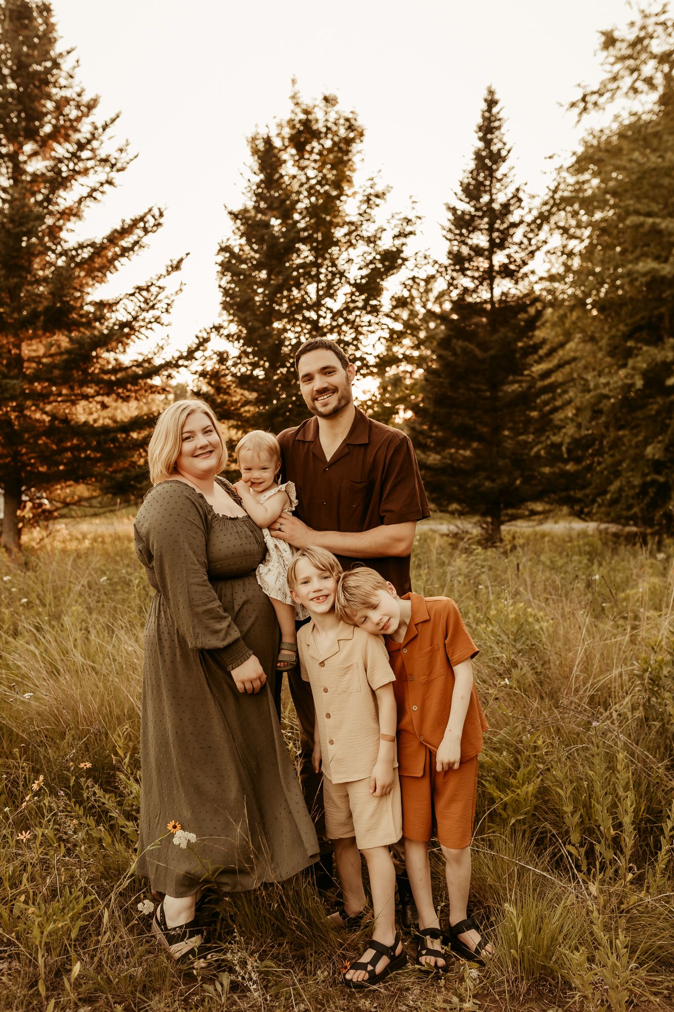 A family poses together in a golden field at sunset with evergreen trees in the background.