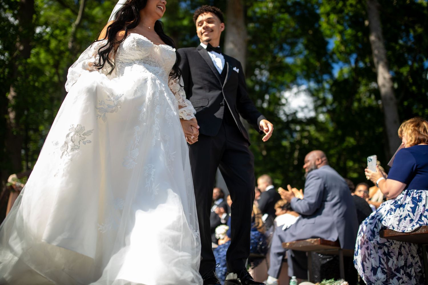 Bride and groom celebrate their wedding with joyful dancing outdoors surrounded by guests on a sunny summer day.