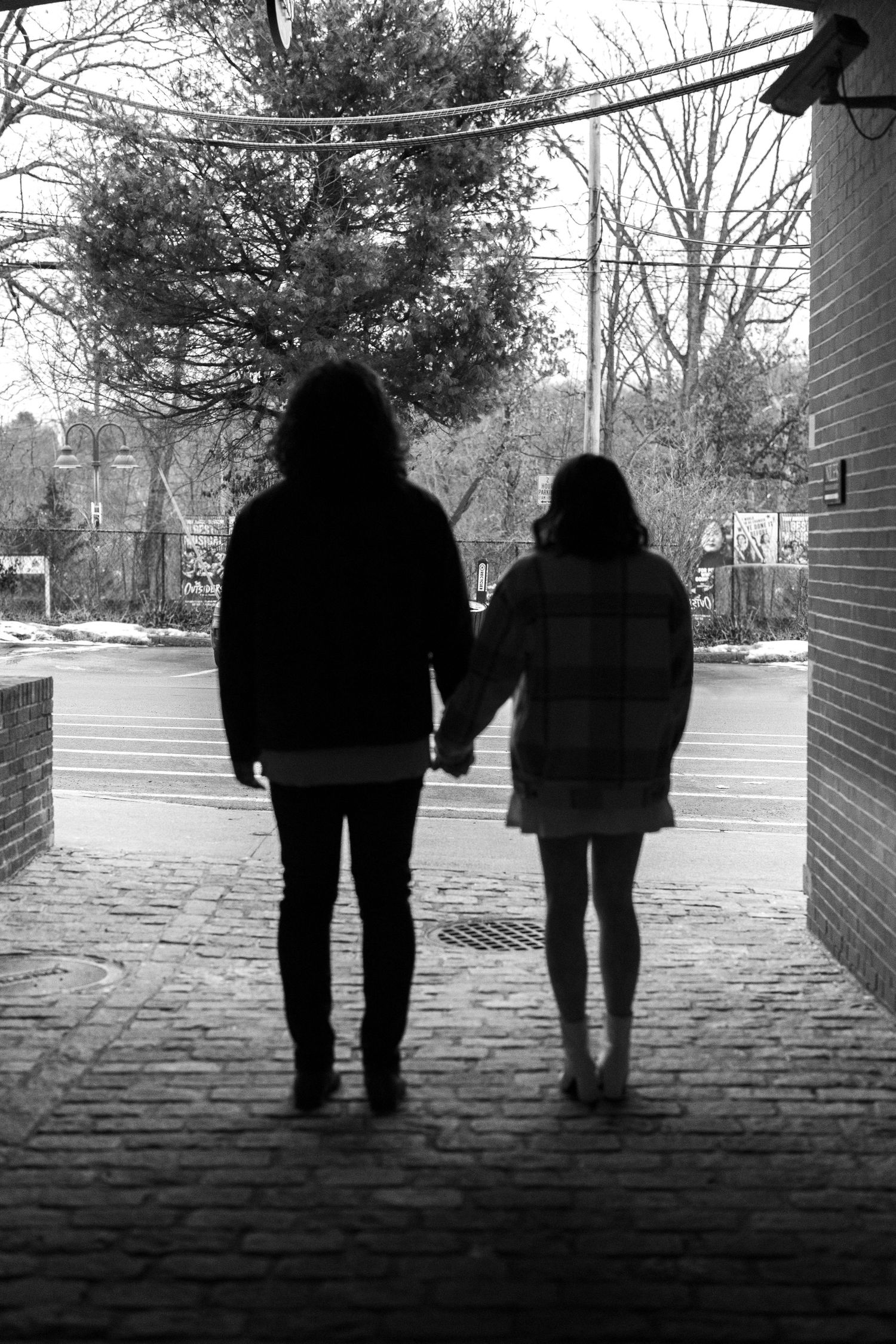 A silhouetted couple holding hands while standing in shadow under a street overhang in black and white.