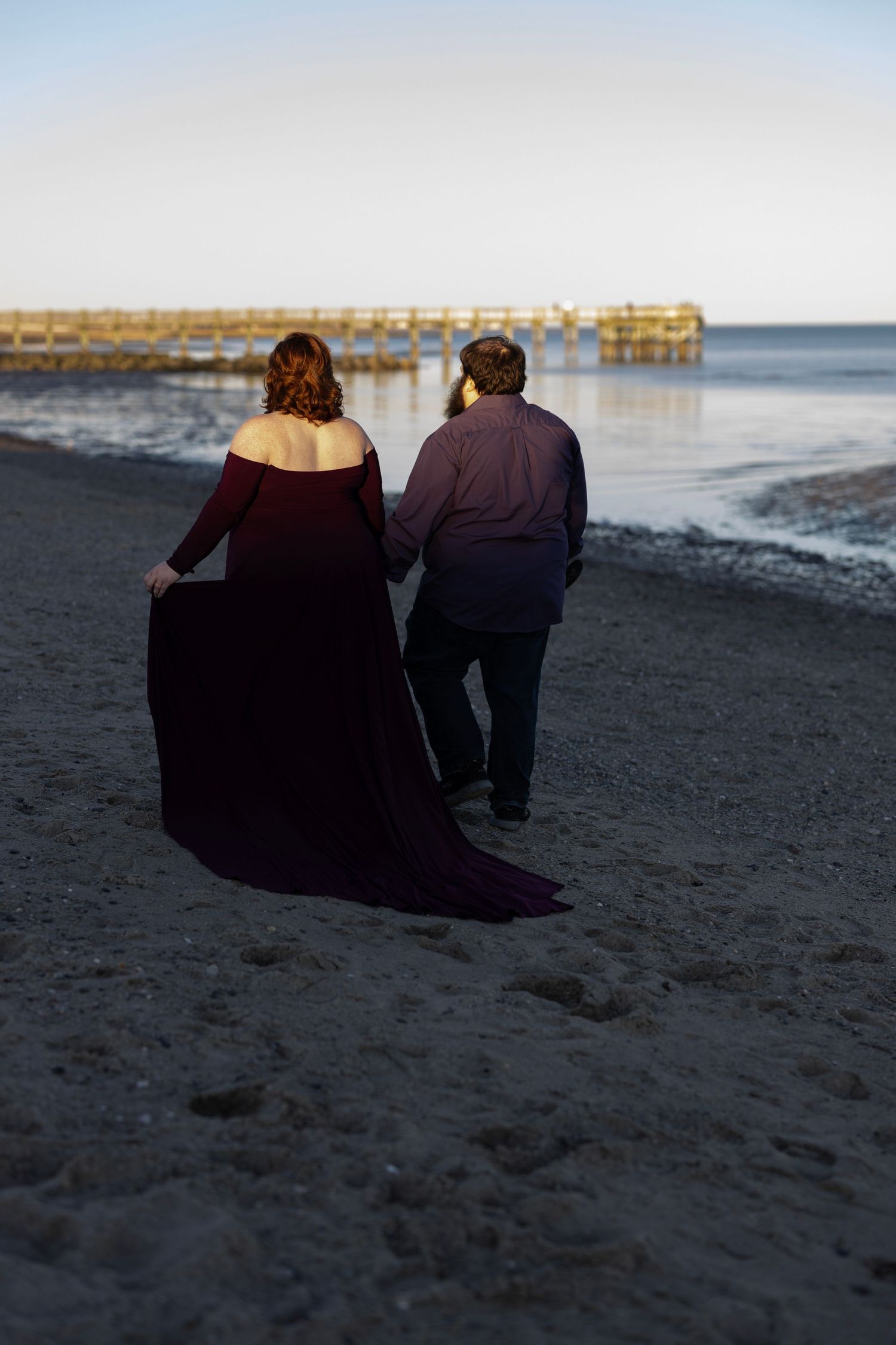 Two people in dark clothing walk along a beach at dusk with a wooden pier in the background.