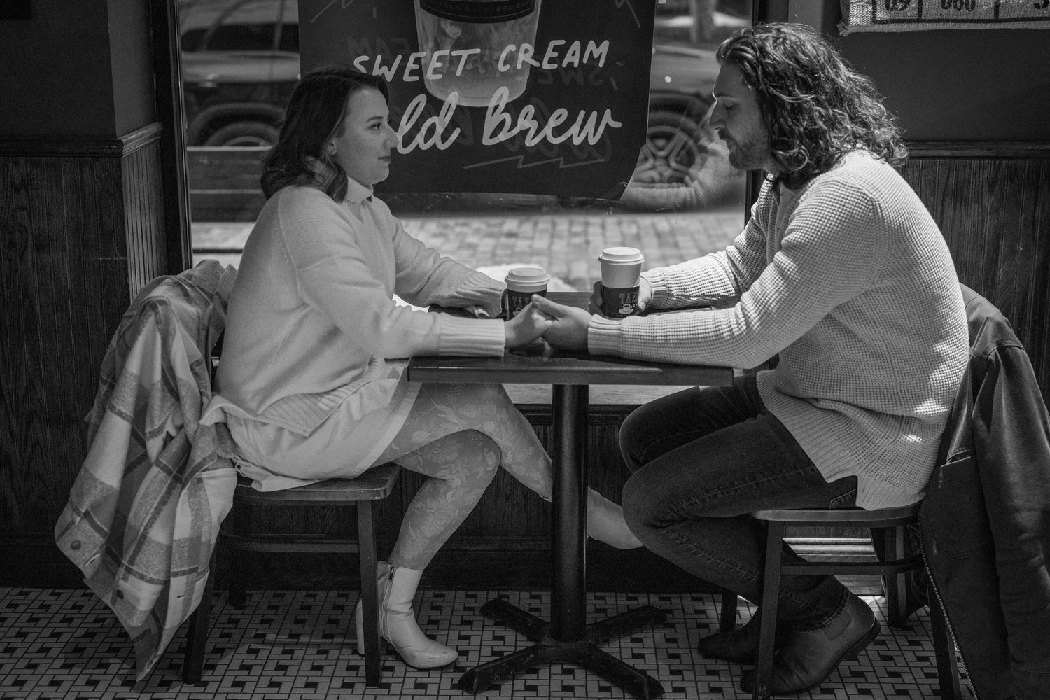 People sit and chat at outdoor cafe tables along a city sidewalk in black and white photography.