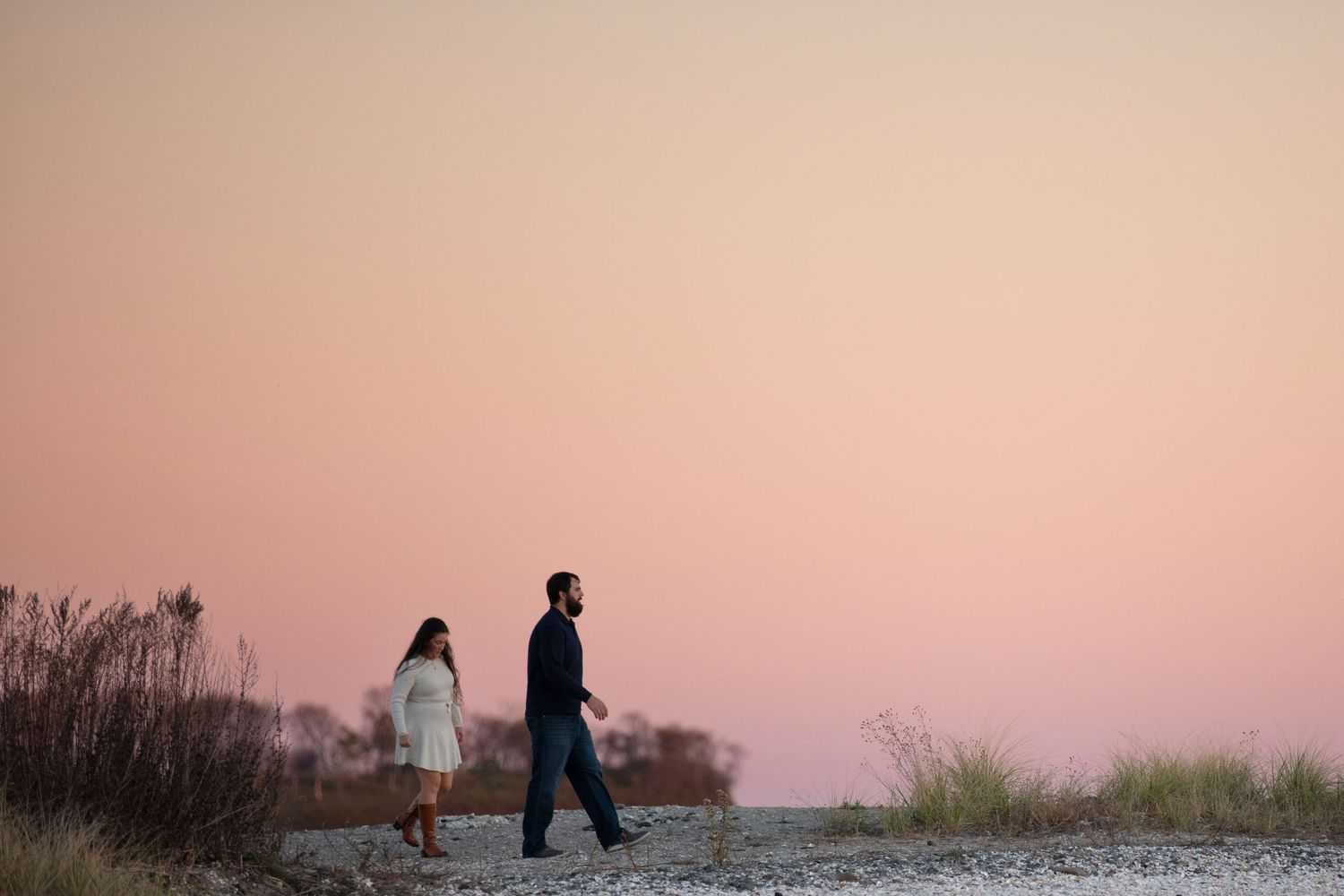 A couple walks along coastal sand dunes during a vibrant pink sunset along the beach.