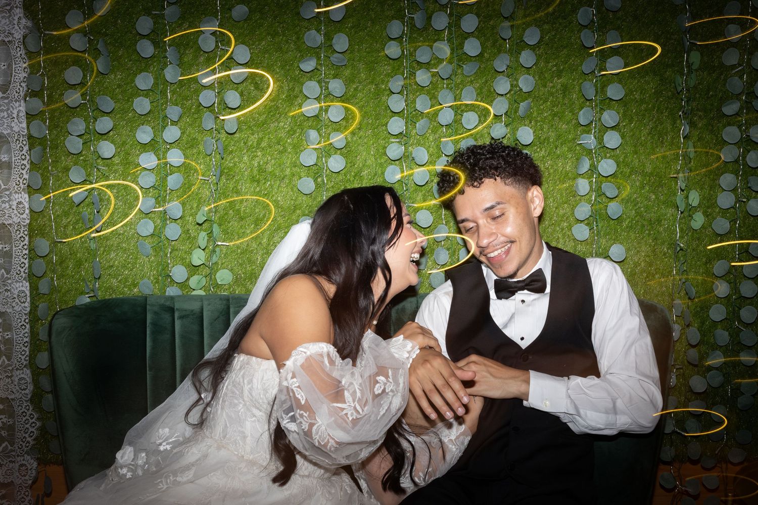 Newlyweds share a joyful moment on an elegant green velvet couch at their wedding reception.