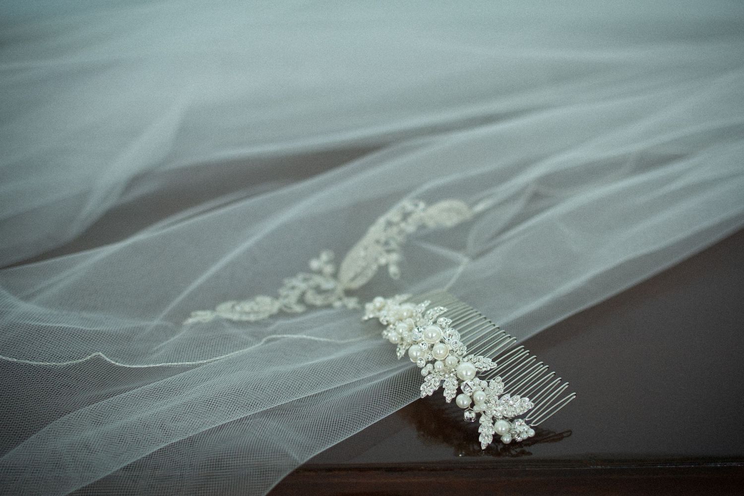 A delicate bridal hair comb with crystal details rests on top of flowing white tulle veil on dark wooden surface.