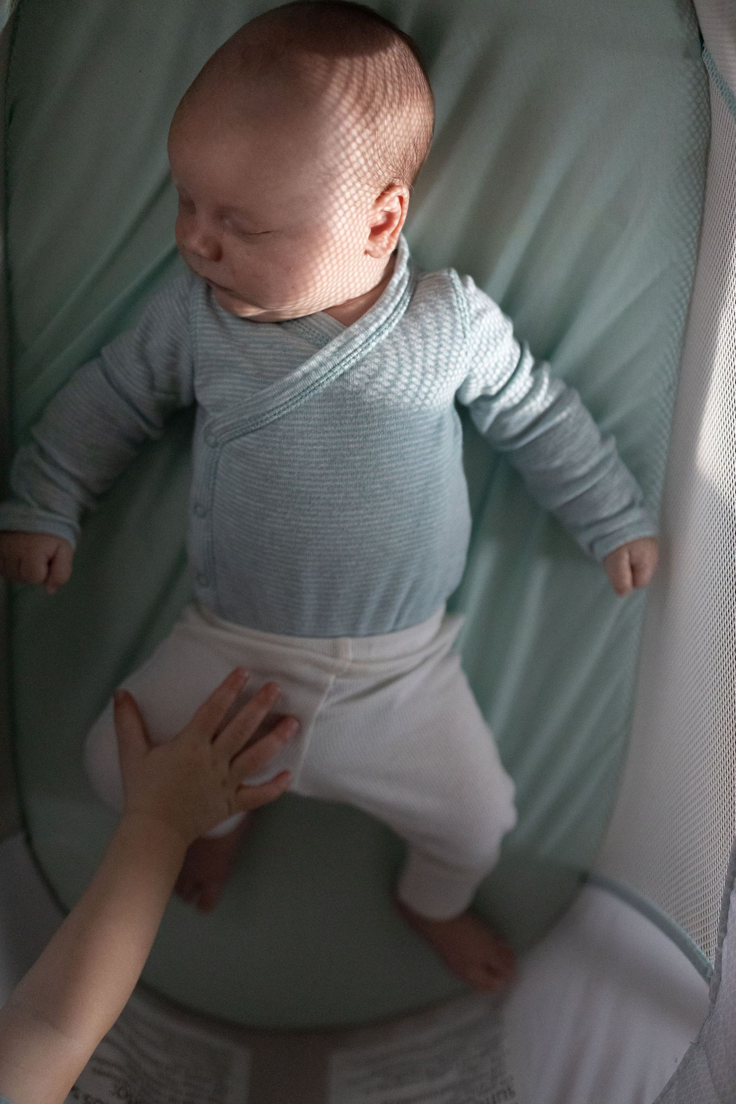 A small baby wearing a gray striped top and white pants rests on a mint green surface while an adult's hand gently touches.