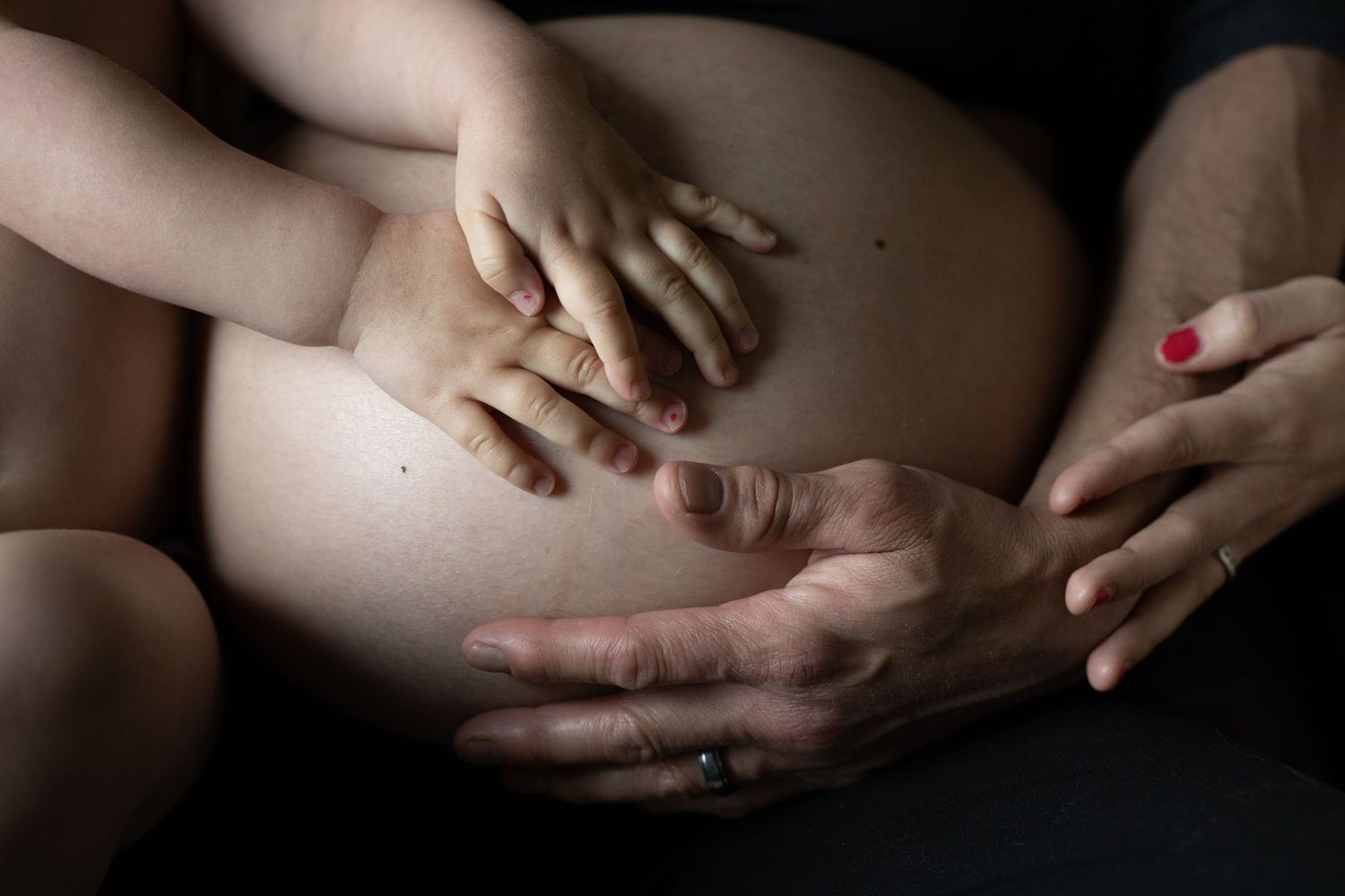 Multiple hands delicately resting on a pregnant belly in dramatic lighting and soft focus.