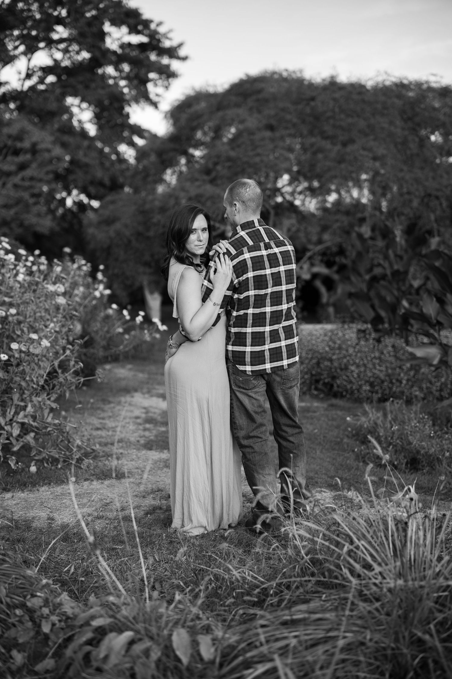 Black and white romantic portrait of a couple embracing in a garden with wildflowers and trees in the background.