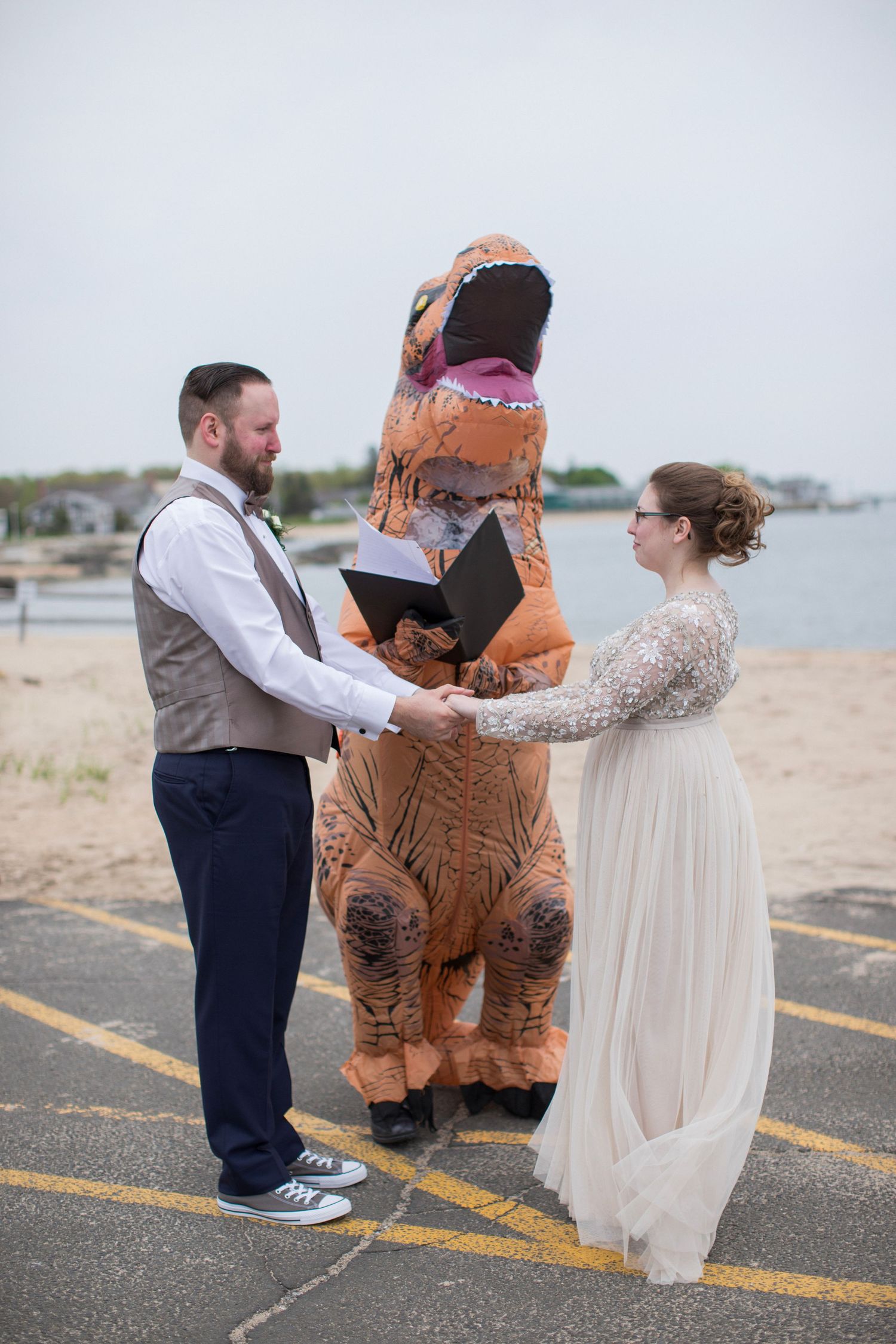 A couple poses for wedding photos with someone in an inflatable T-Rex costume on a beach parking lot in a playful scene.
