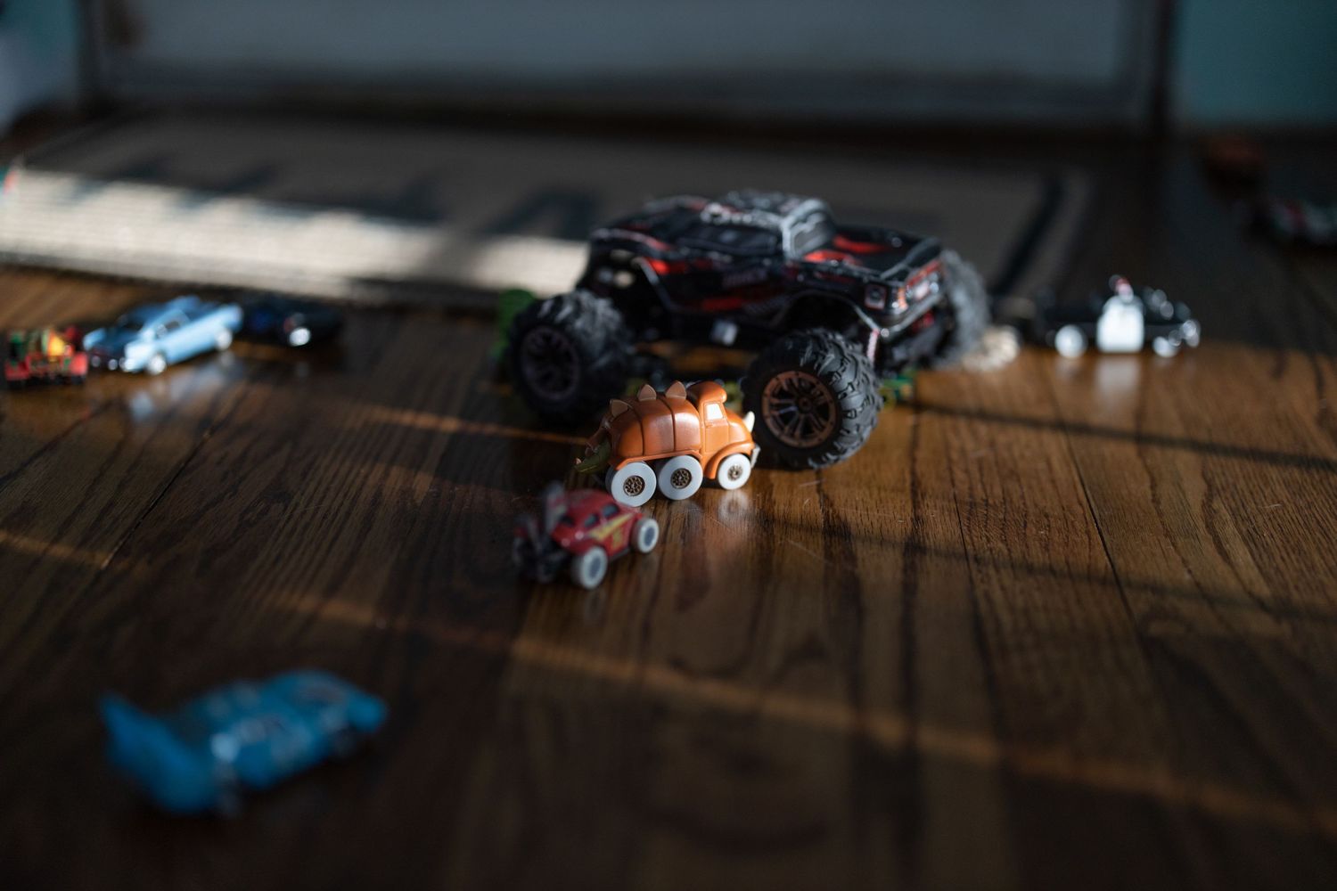 Small toy monster truck with oversized tires sits on wooden floor with blurry toy cars scattered around.
