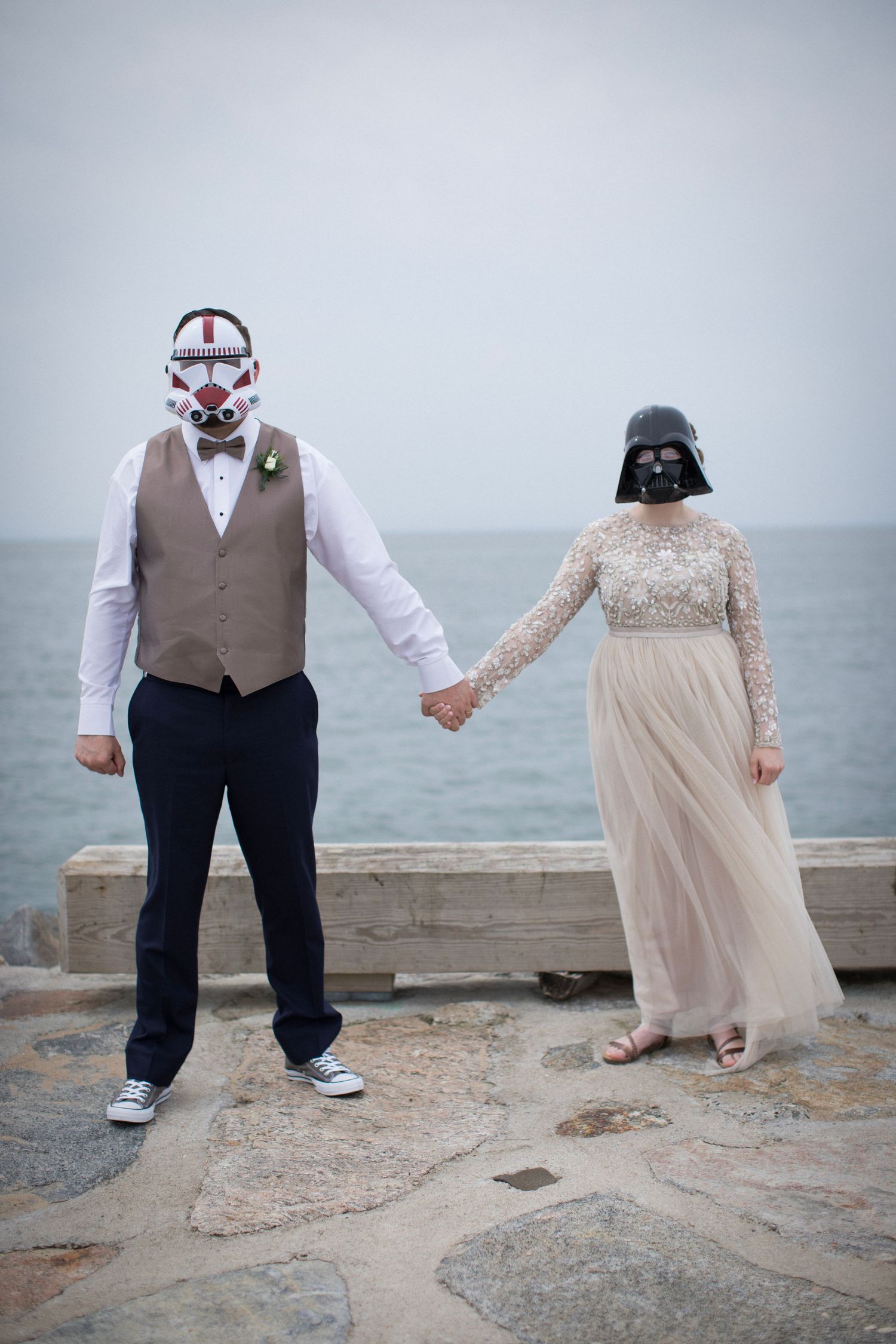 A couple wearing Star Wars inspired masks holds hands on a rocky beach in formal wedding attire.