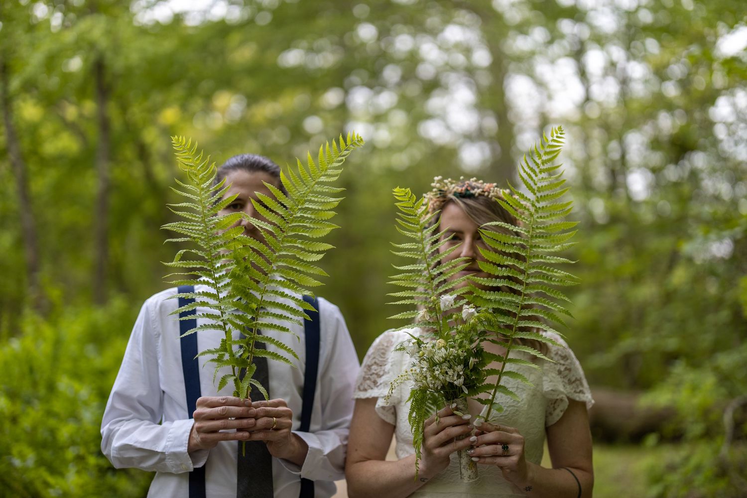 Two people in casual attire stand outdoors holding green fern leaves in front of their faces in a playful nature scene.