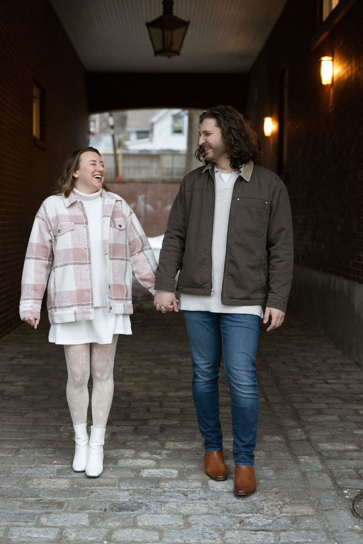 A couple holding hands while walking through a dimly lit brick tunnel, wearing casual winter outfits.