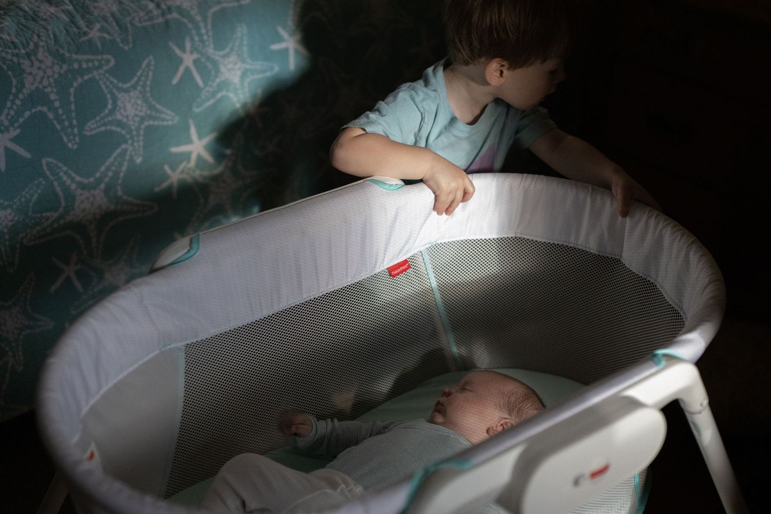 A young child shines a small light over a baby sleeping in a white bassinet while standing in the dark.