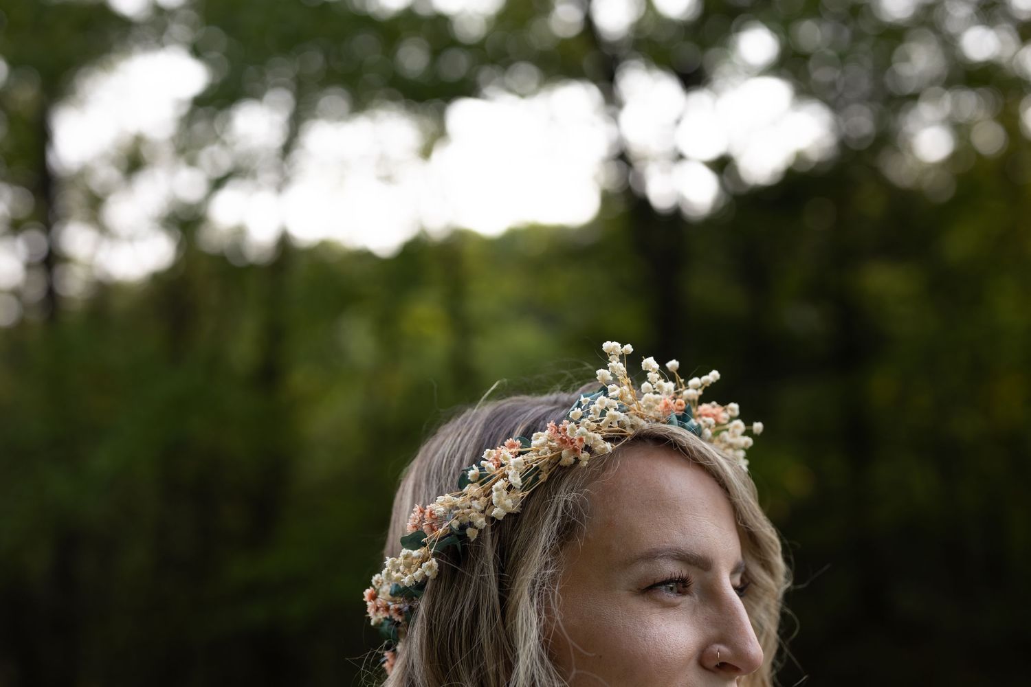 A delicate floral crown adorns someone gazing to the side in a dreamy outdoor setting with bokeh background.