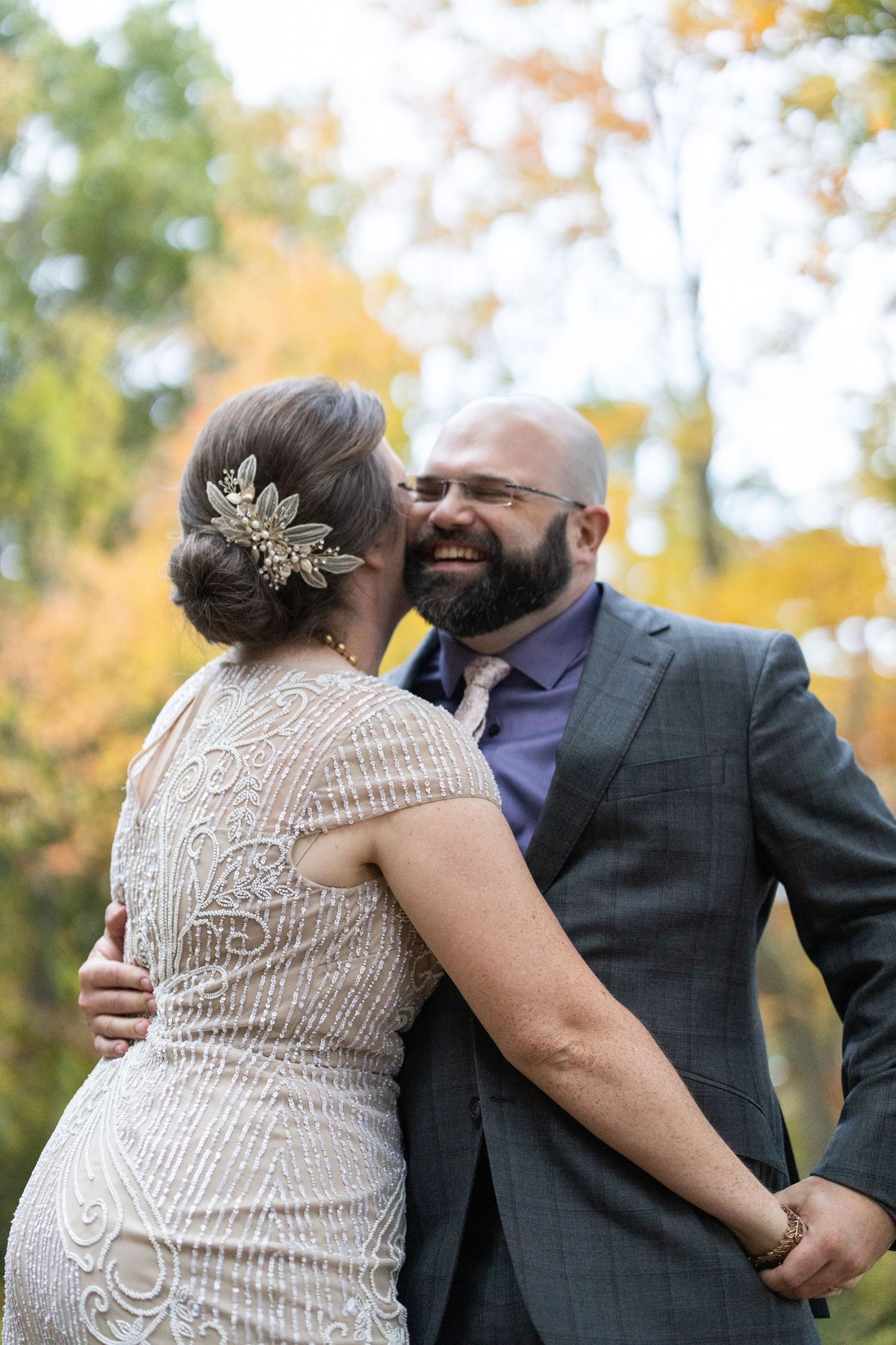 A joyful wedding couple embraces outdoors during autumn, with the bride wearing a detailed lace gown and groom in grey suit.