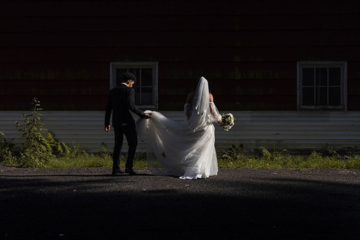 A wedding couple walks across a sunlit outdoor path while a photographer captures their special moment.