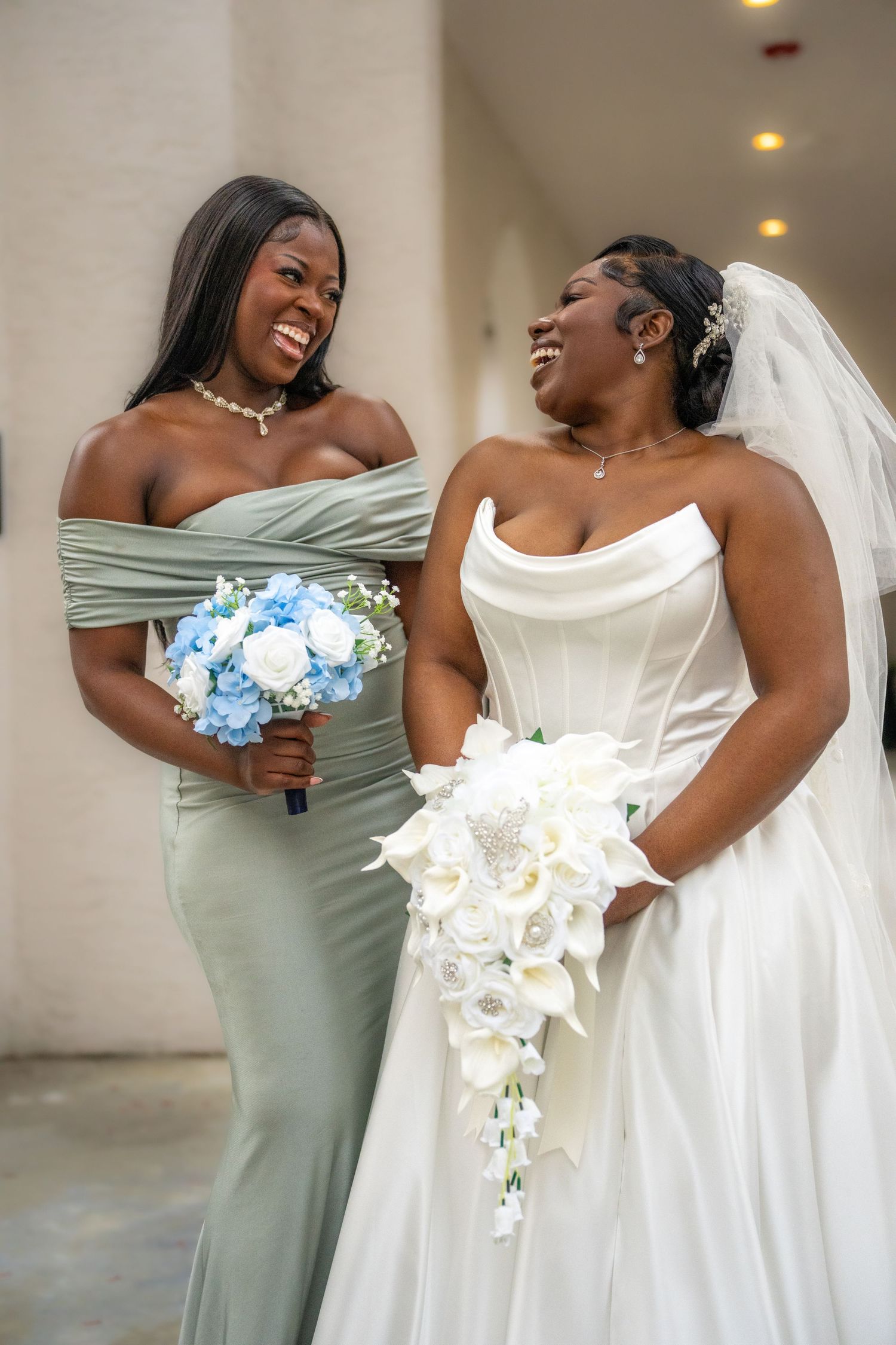 A bridesmaid in sage green holds blue flowers next to the bride with a cascading white bouquet.