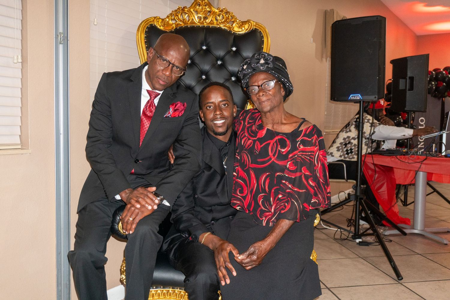 Three people pose together on an ornate gold chair at a formal event with red decor visible in the background.