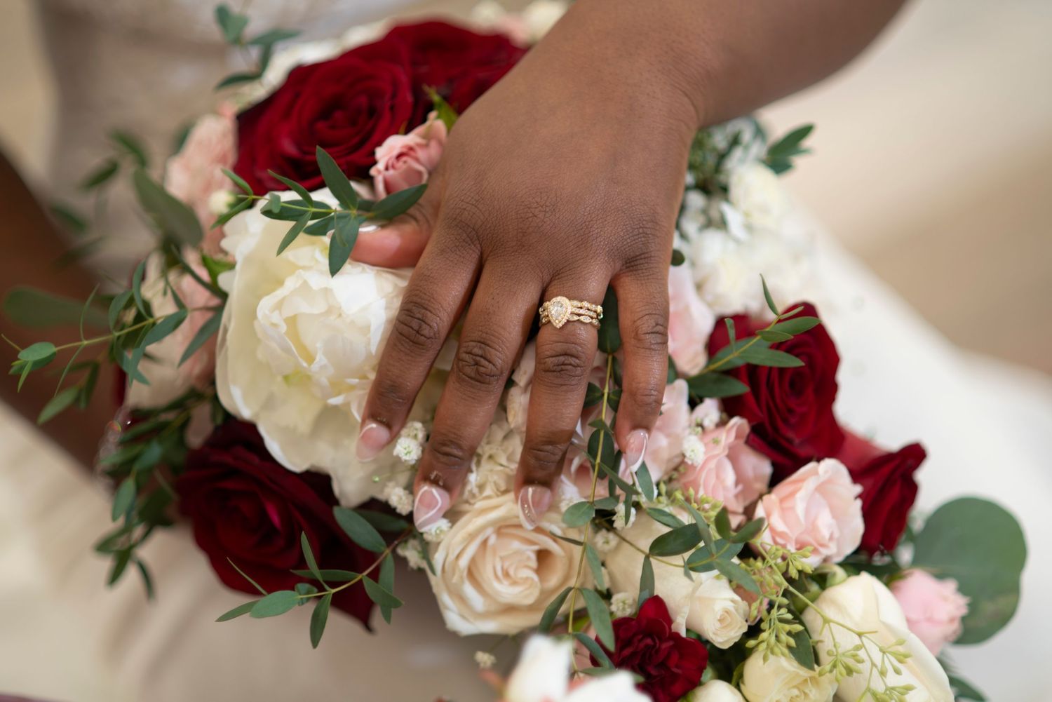 A hand wearing a wedding ring rests on a bridal bouquet of red and white roses with greenery.