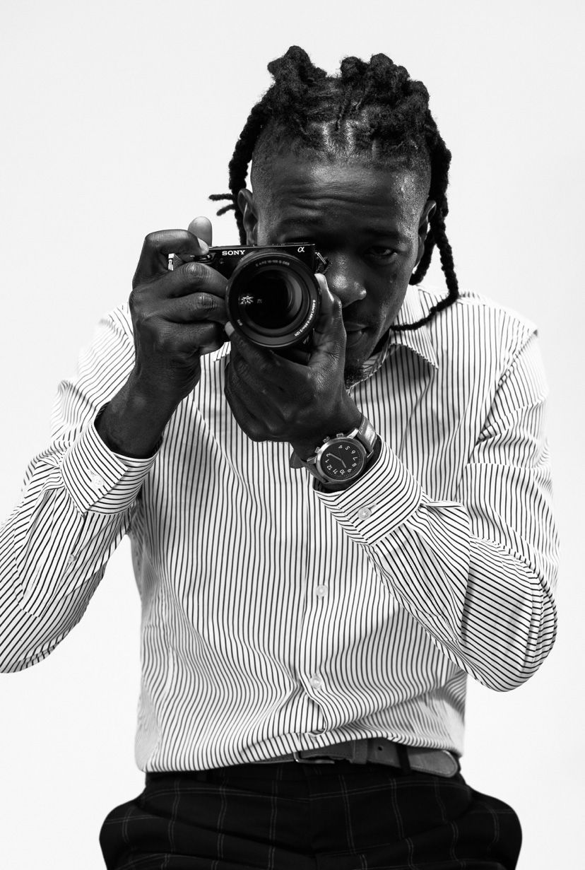 A photographer wearing a striped button-down shirt and watch holds up a professional camera in black and white portrait style.